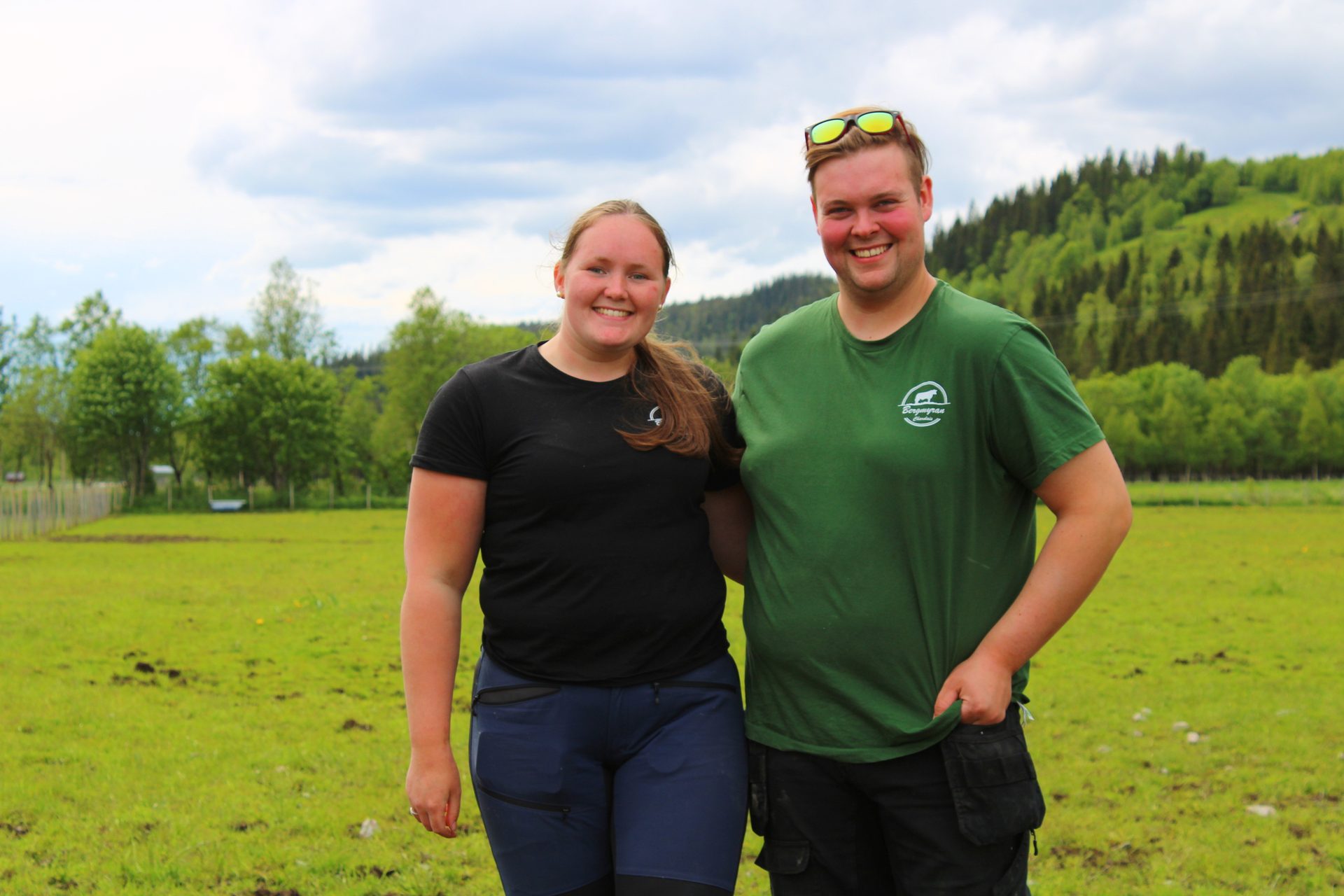 Smiling man and woman in a green field with wooded hills.