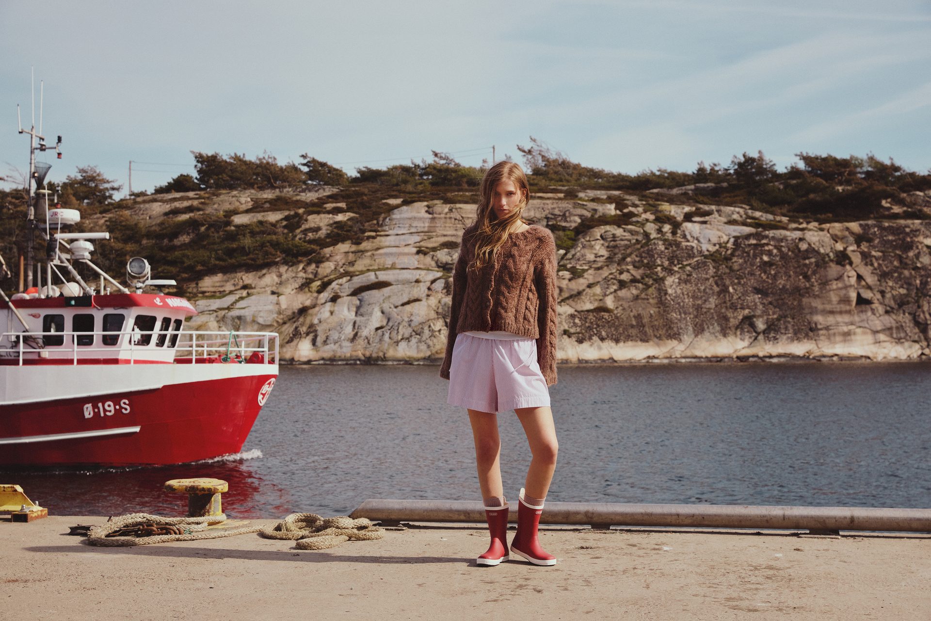 Young woman in sweater & red boots on a pier, with a red fishing boat and rocky cliffs.