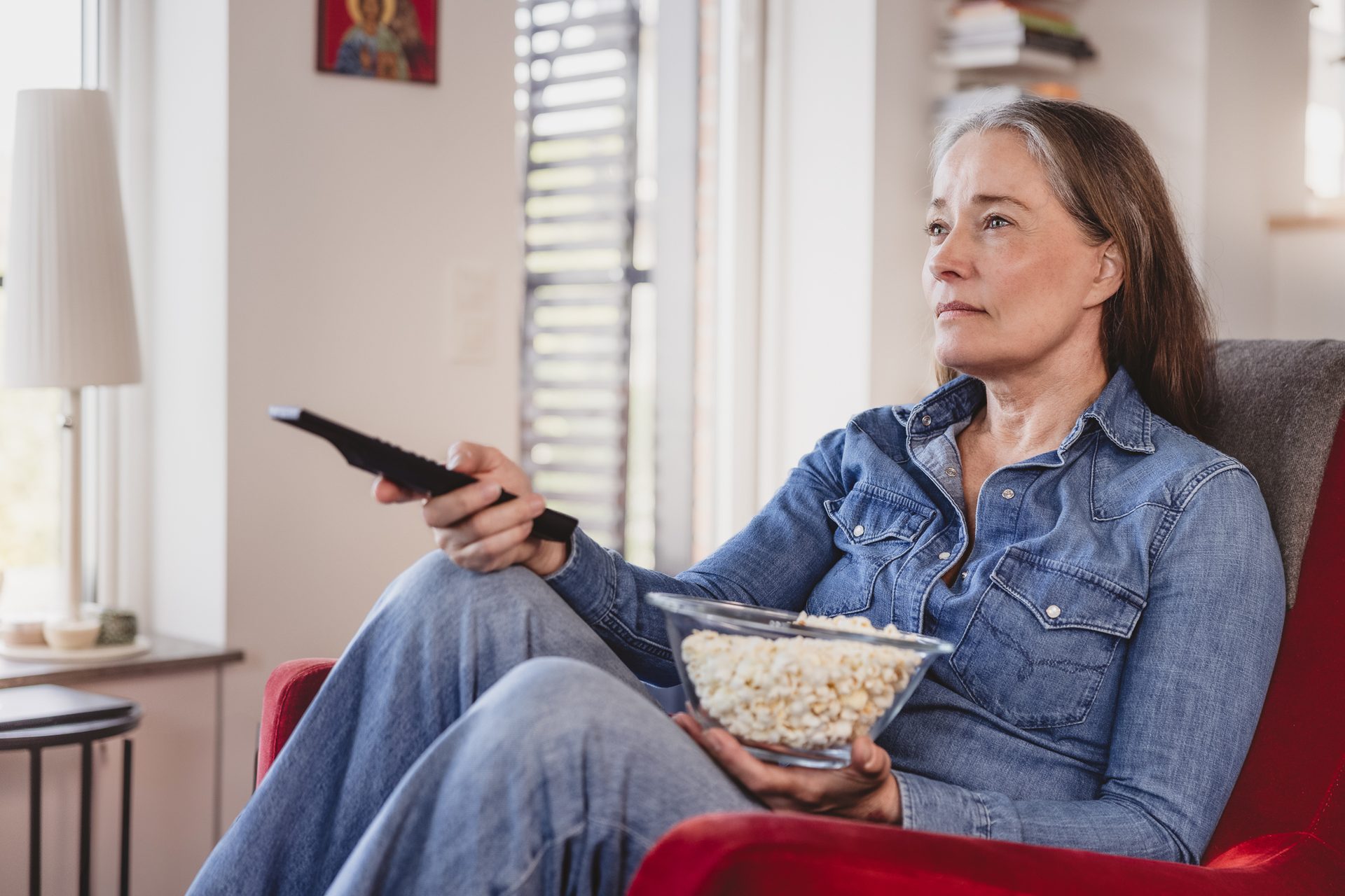 An older woman sits in a chair with popcorn and a remote, watching TV.