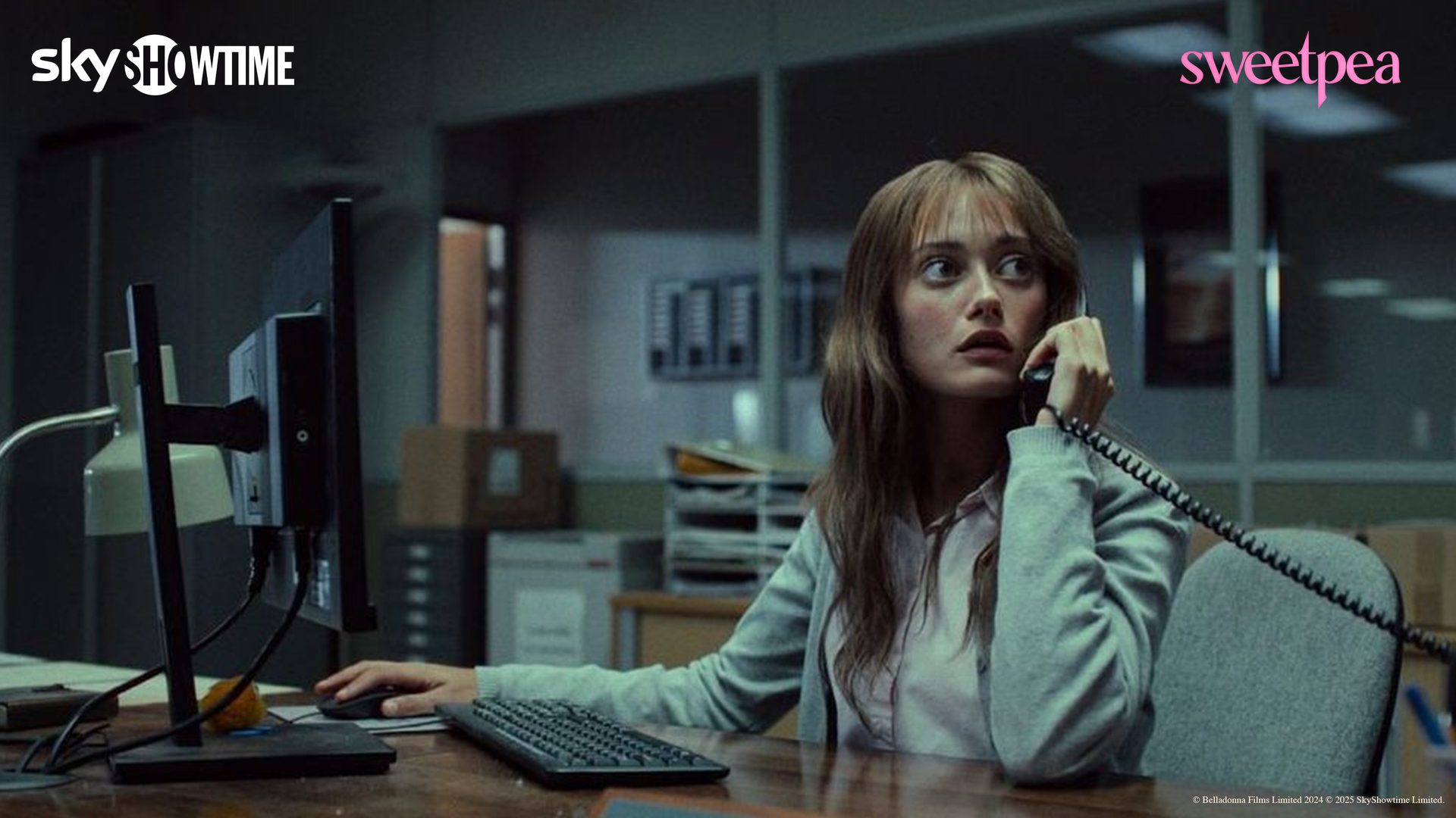 A young woman with bangs at an office desk, talking on a phone with a worried expression.