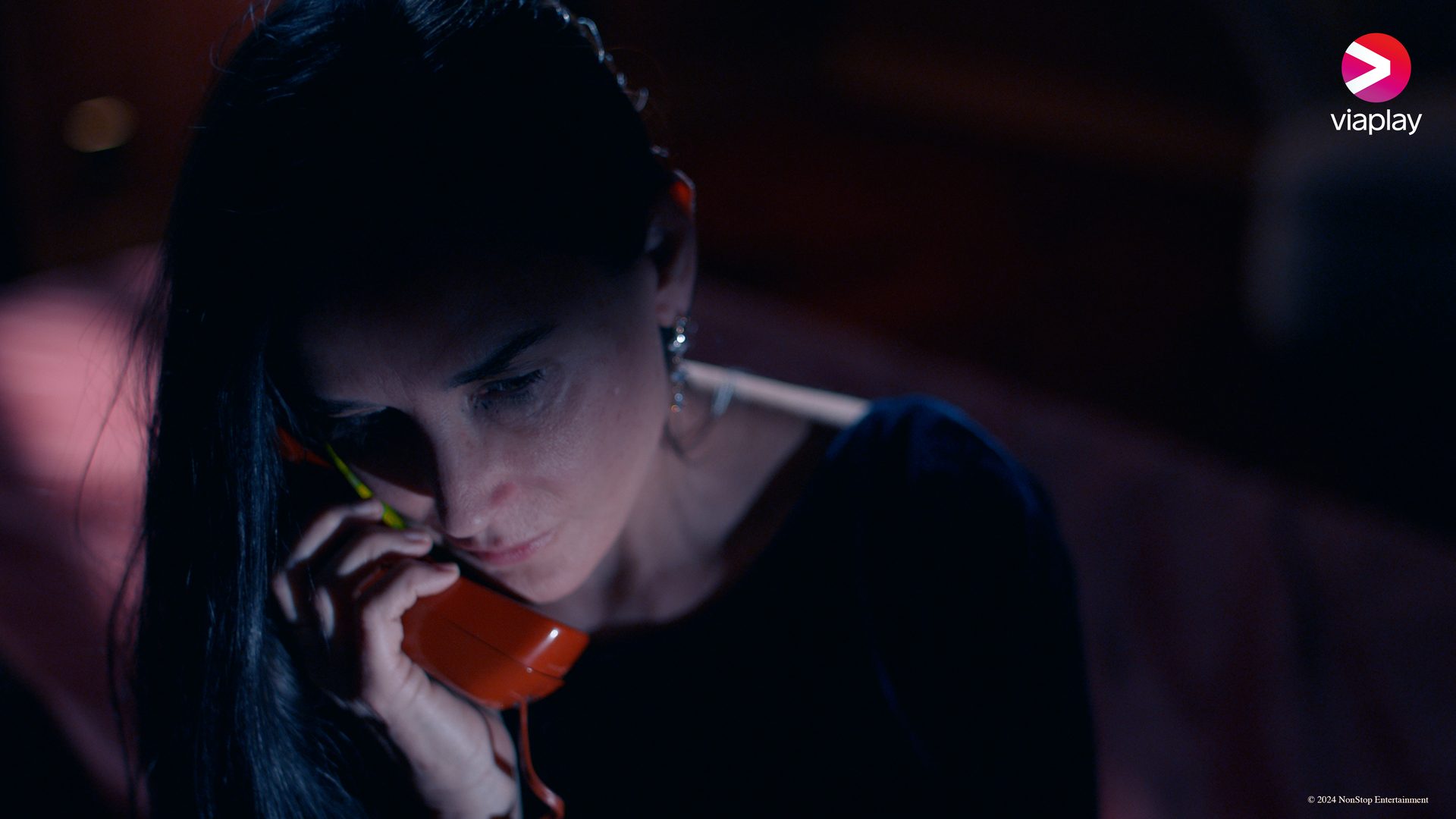 A woman in dim light holds a red telephone receiver to her ear, looking down thoughtfully.