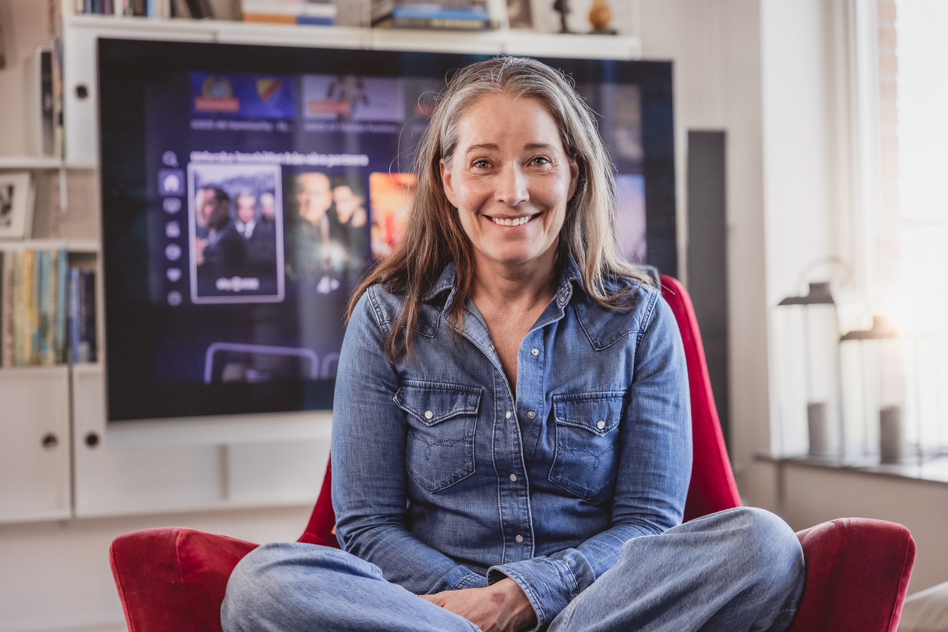Smiling woman in denim sits in red armchair, TV behind her.