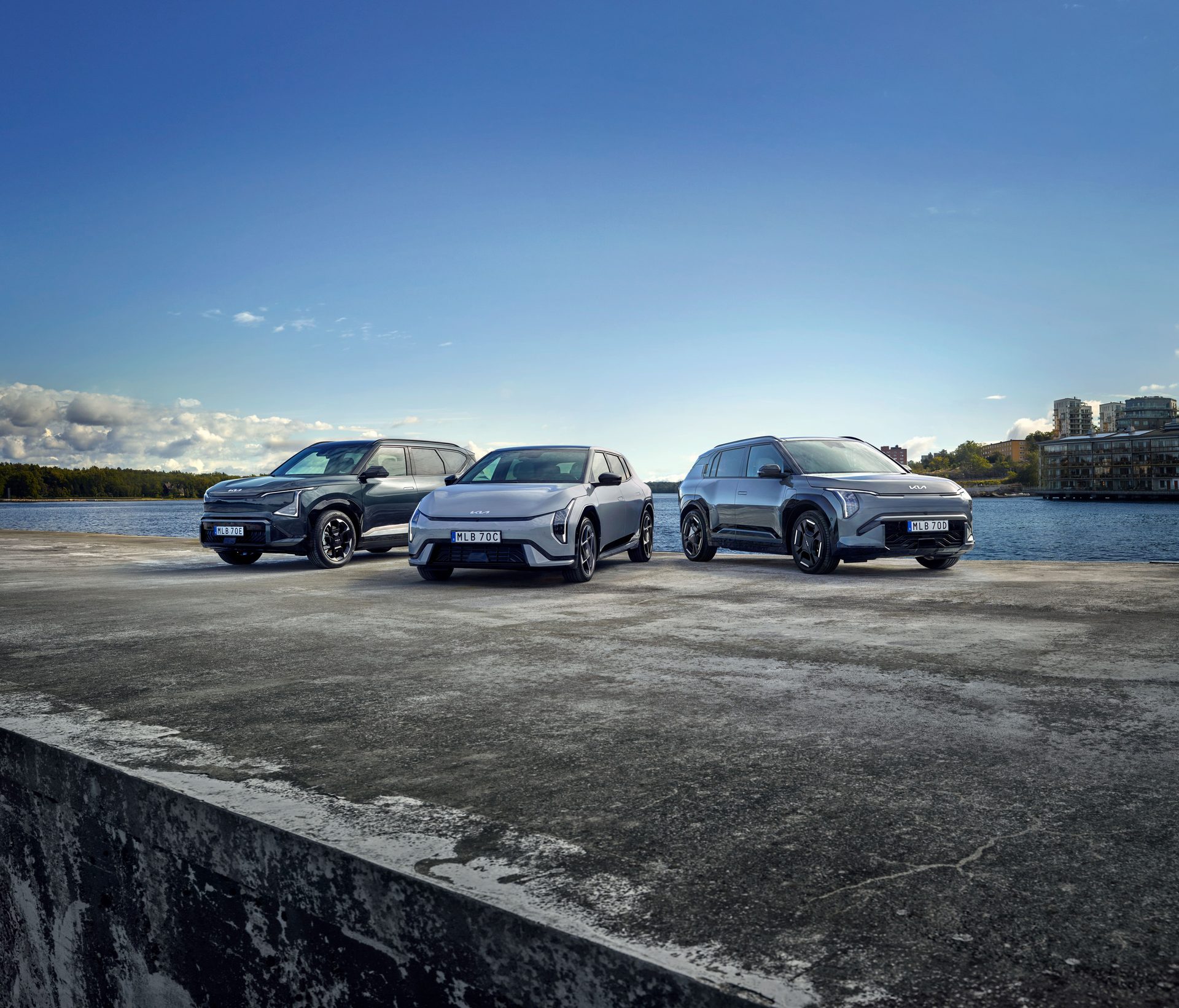 Three modern cars on a concrete pier by the water, with a cityscape and trees under a blue sky.