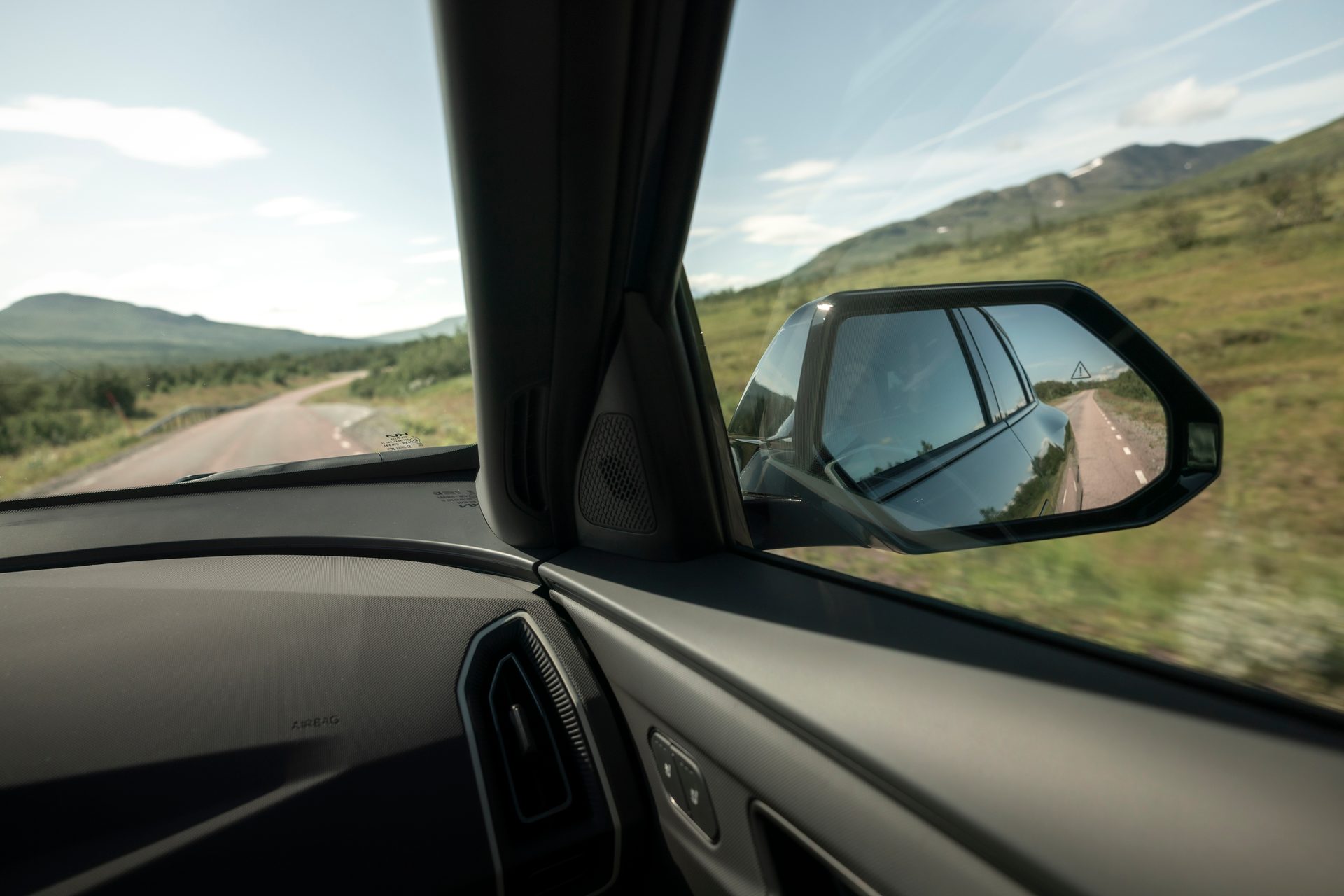 View from inside a car of a winding mountain road, with the side mirror reflecting the road behind.