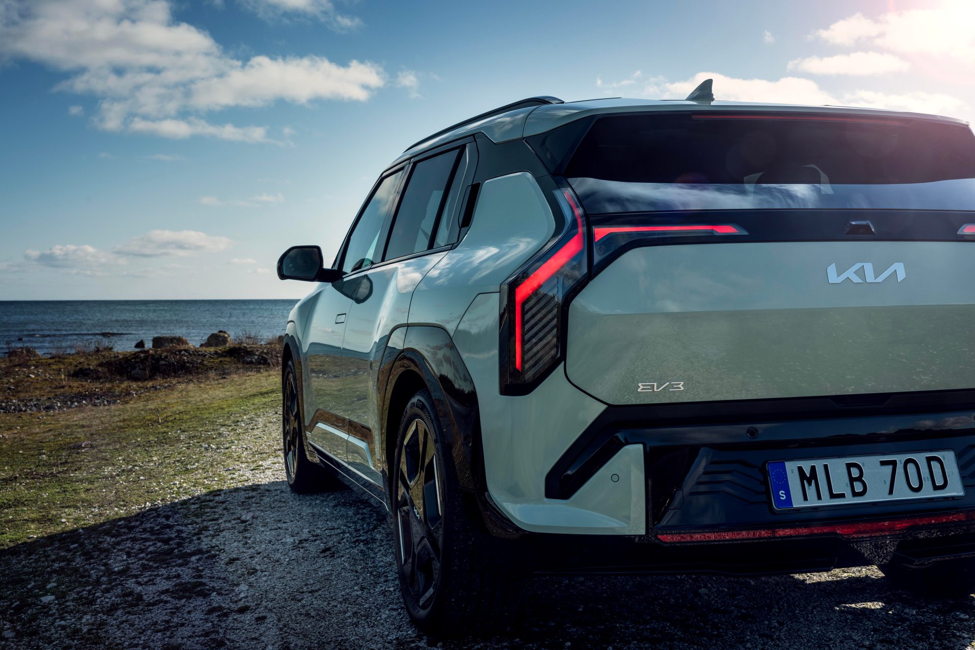 Light green Kia EV3 SUV, rear-side view, parked by the sea under a cloudy sky.
