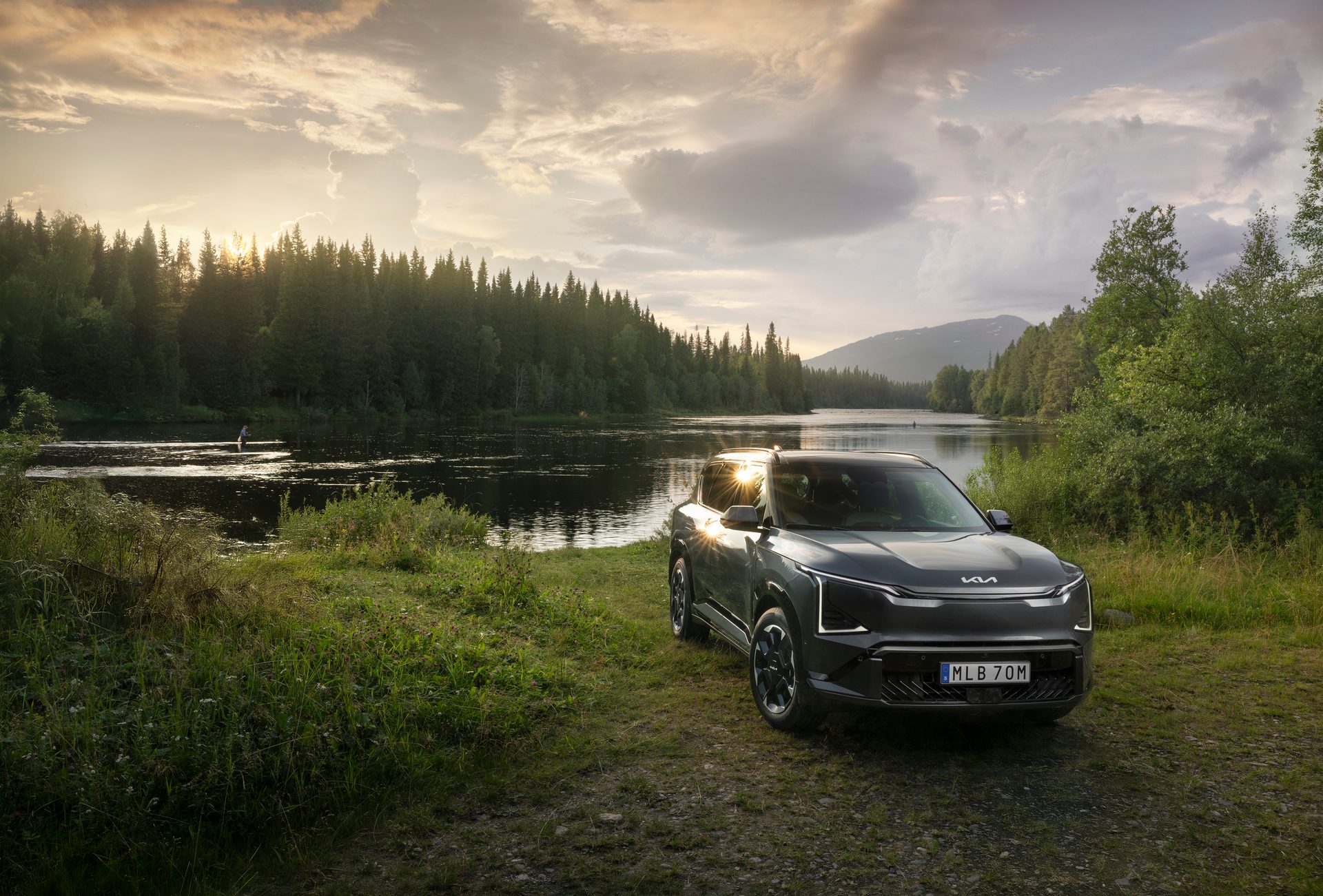 Kia EV9 electric SUV parked by a forest lake at sunset.