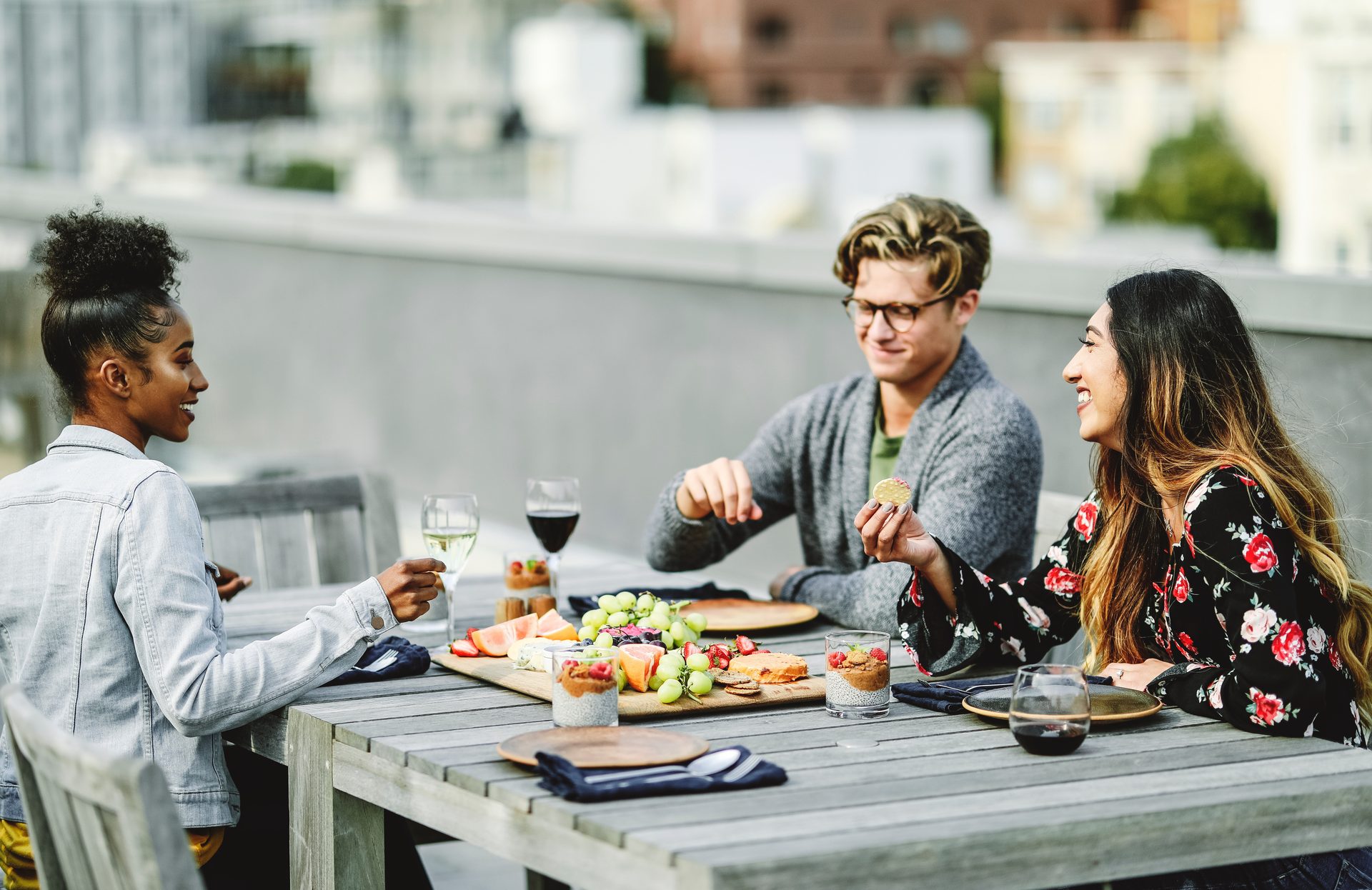 Three diverse friends enjoy wine and a charcuterie board on a rooftop patio, laughing and socializing.