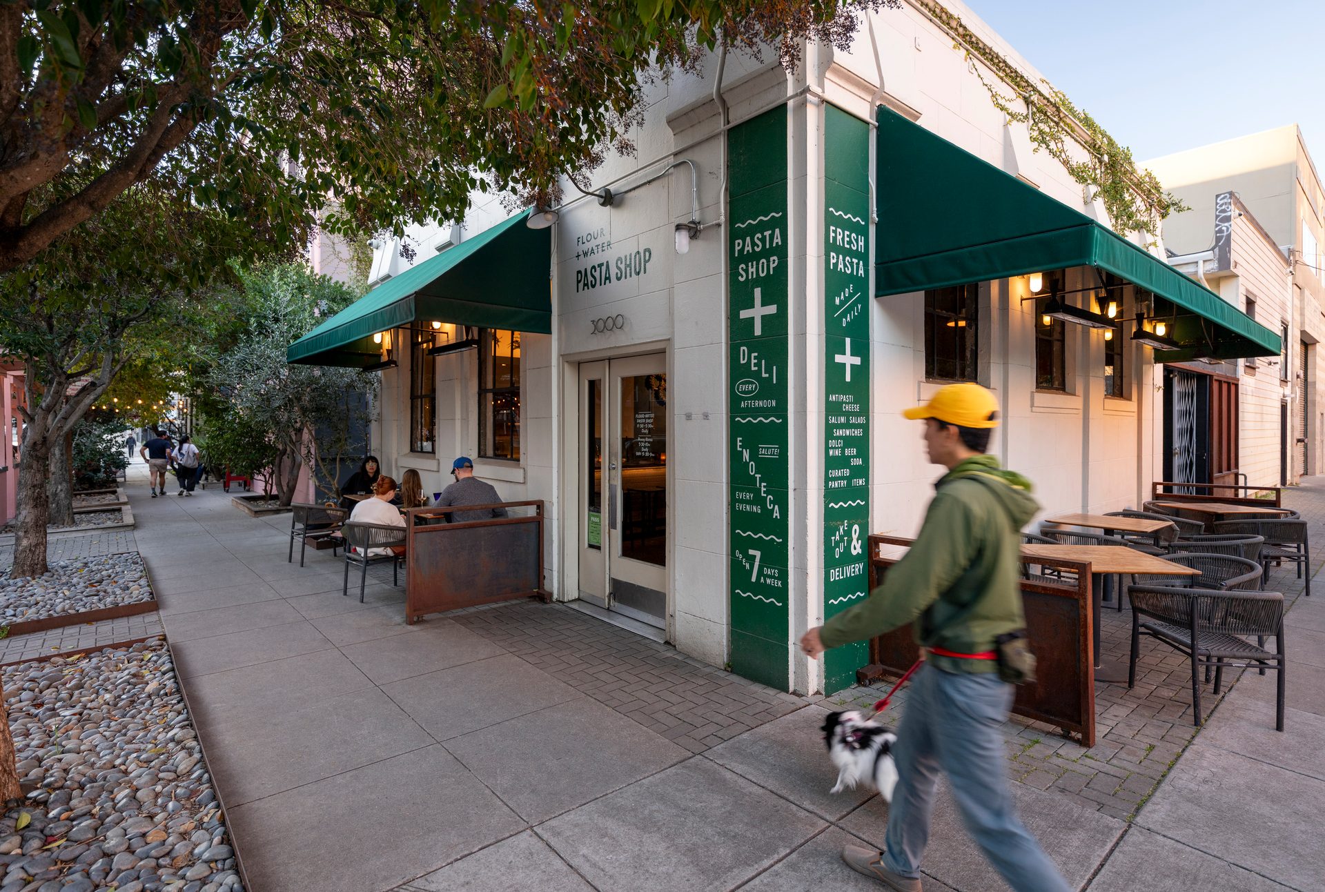 A Flour + Water Pasta Shop with green awnings and outdoor dining, as a man walks a dog.