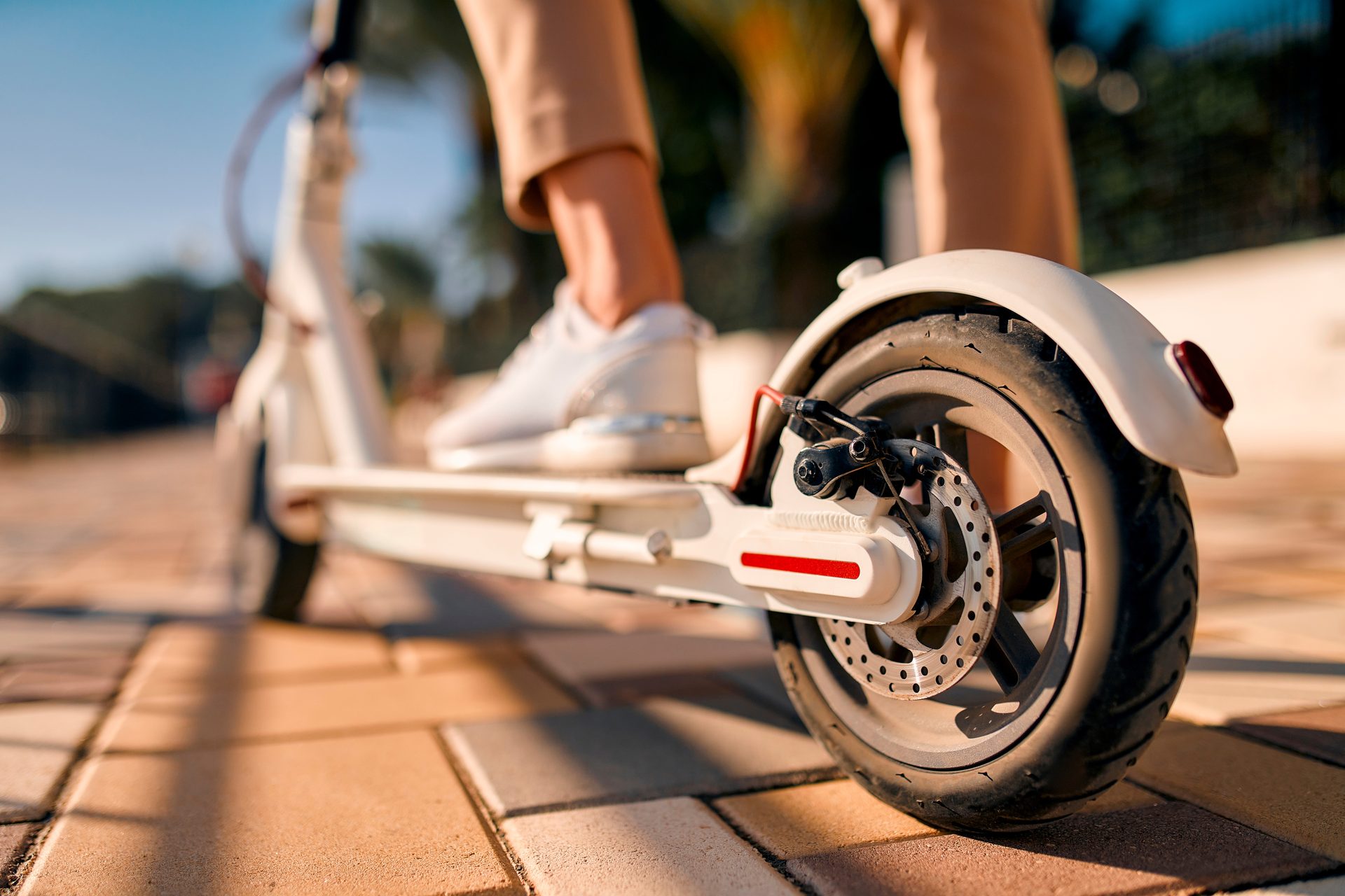 Close-up of a person's feet in white sneakers on an electric scooter, focusing on the rear wheel and brake.