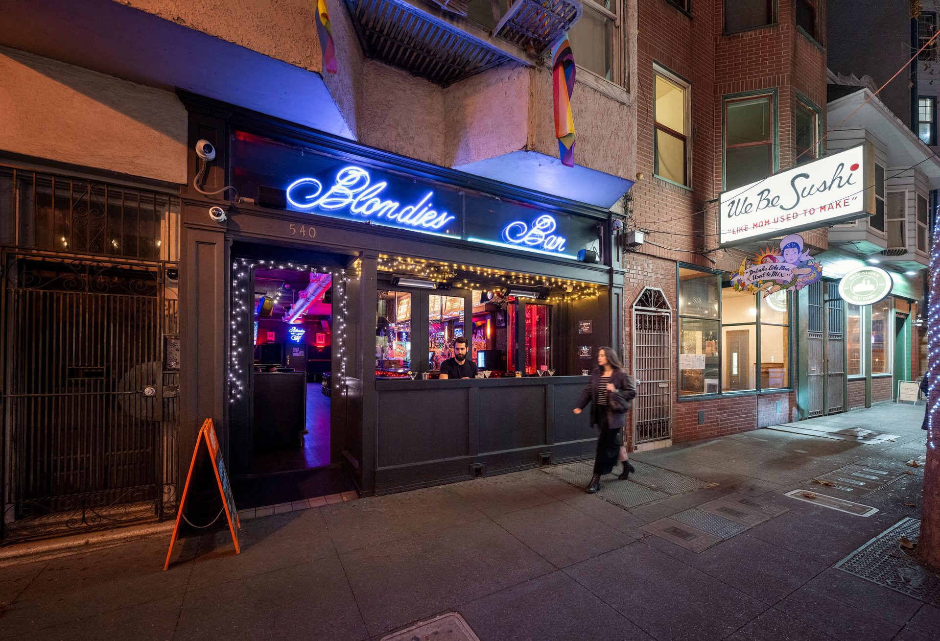 Night shot of Blondies Bar with neon signs, a person walking, and We Be Sushi next door.