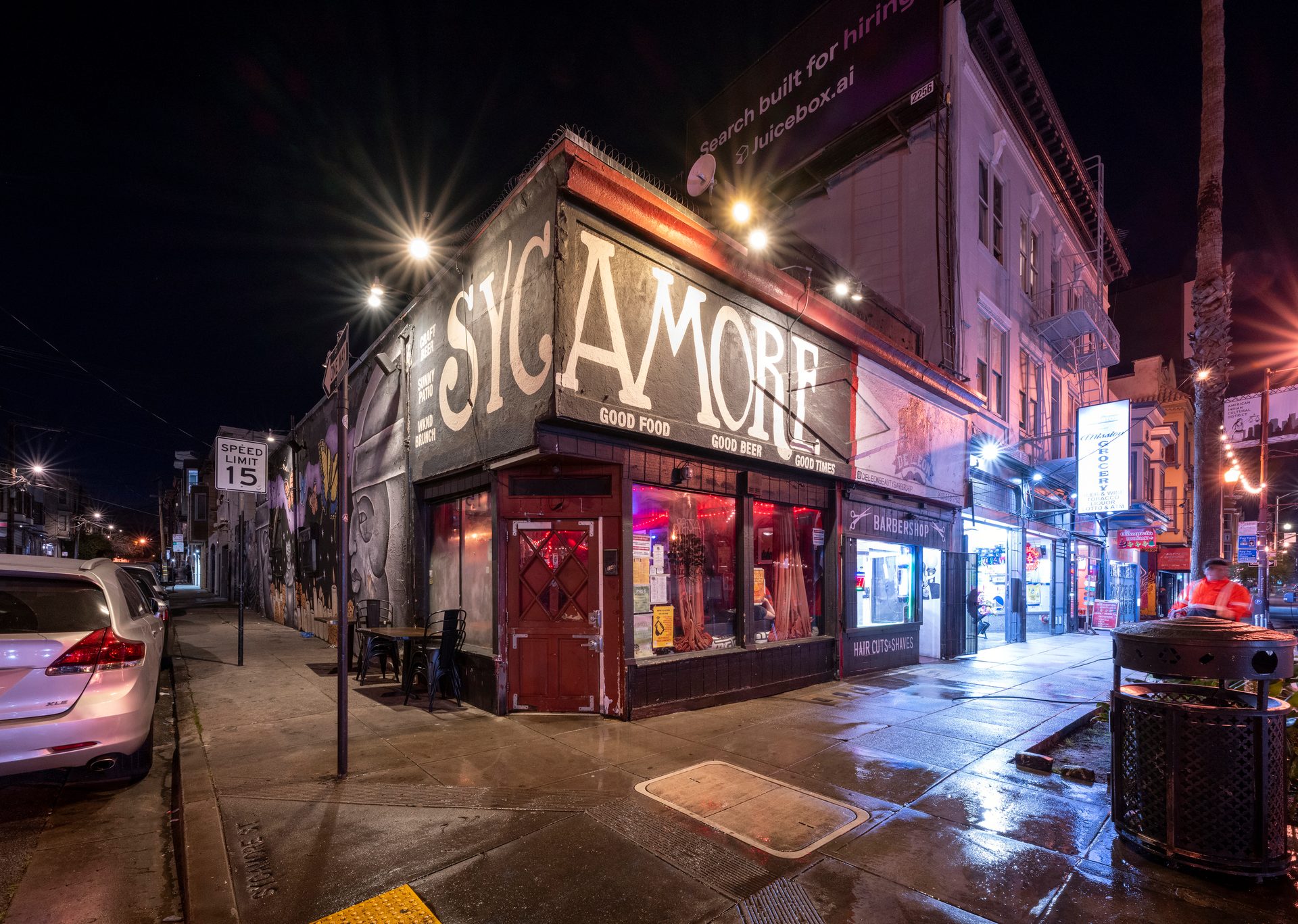 Night view of "The Sycamore" bar/restaurant on a city corner with a barbershop, street lights, and wet pavement.