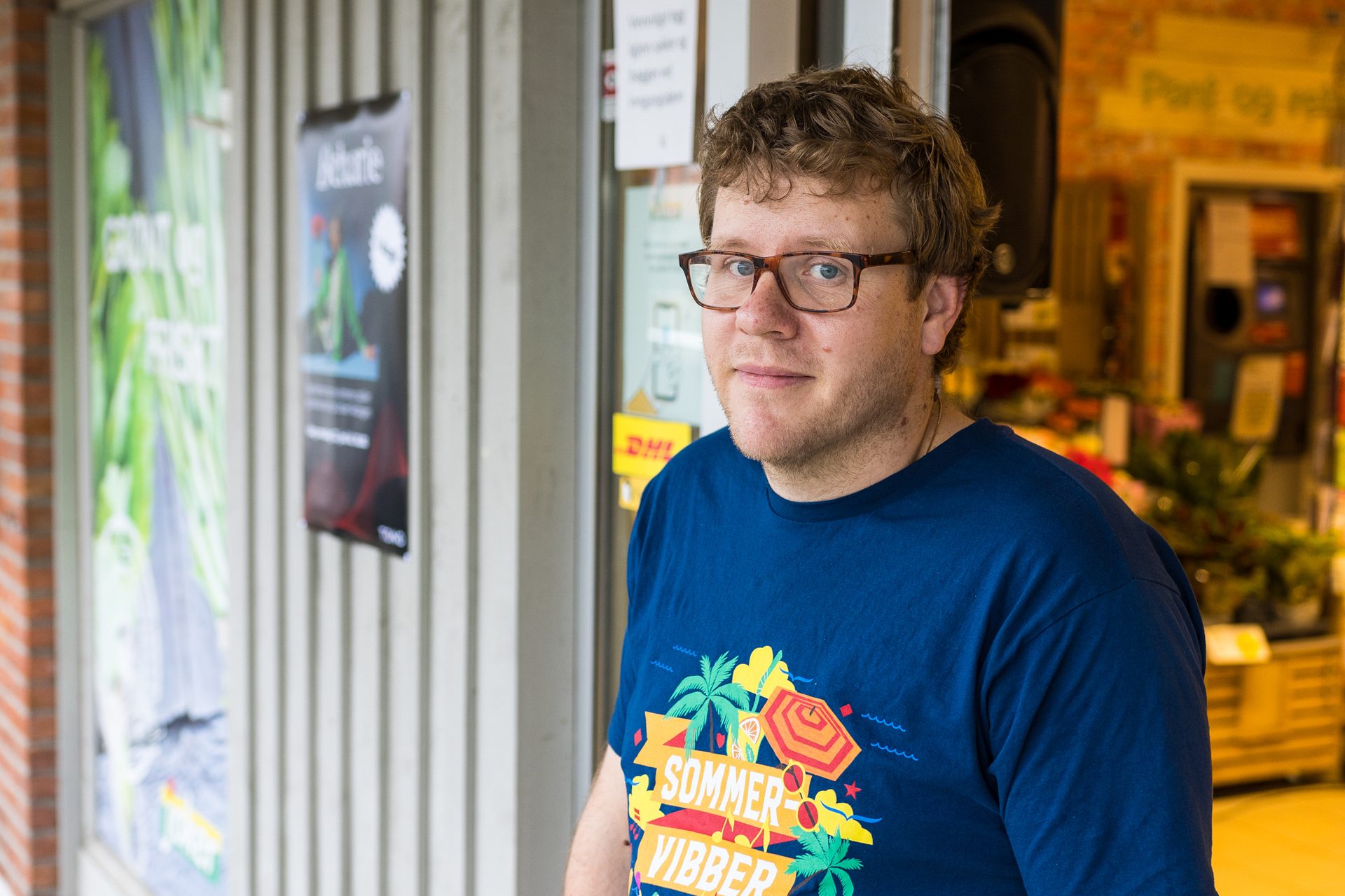 Portrett av en mann med m&#xF8;rkt blondt h&#xE5;r og briller st&#xE5;r i inngangen av en matbutikk