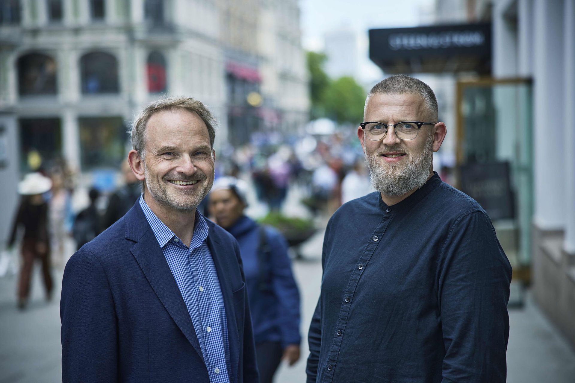 Two smiling men on a city street with a blurred urban background.