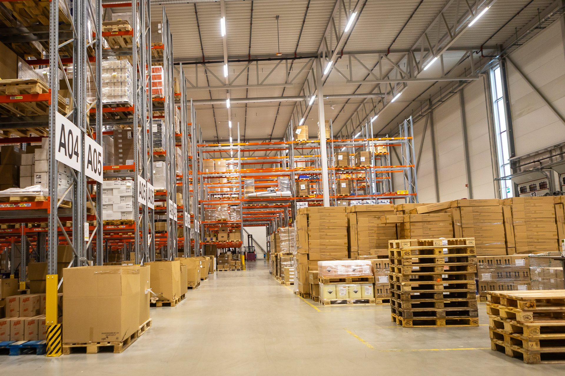 A large, bright warehouse interior with tall industrial shelving units packed with cardboard boxes and pallets.