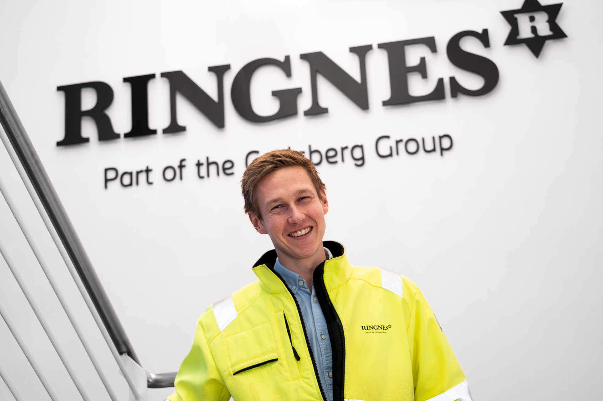 Smiling man in yellow jacket stands by a prominent 'RINGNES' sign.
