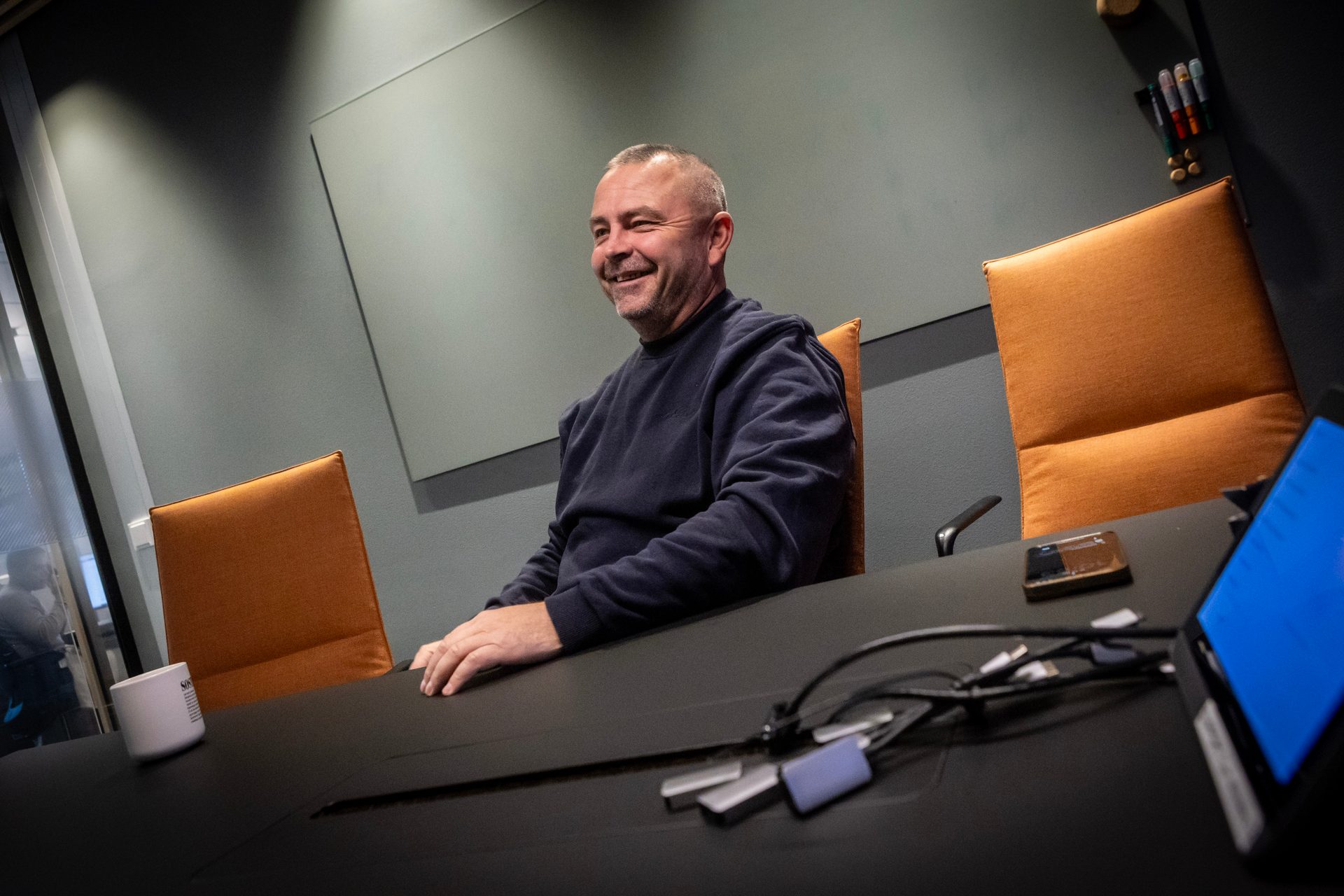 A smiling man in a dark sweater sits at a conference table, looking left in a modern room.