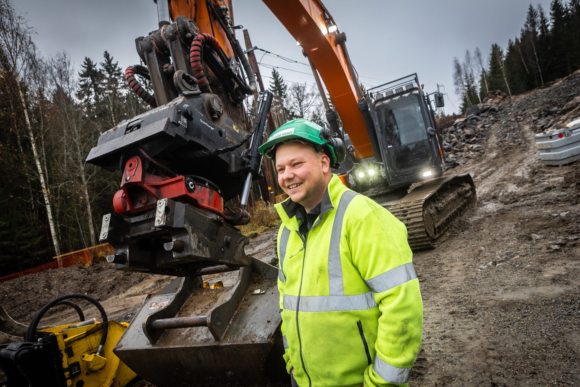 Smiling construction worker in a hi-vis jacket and hard hat, with an excavator at a muddy site.