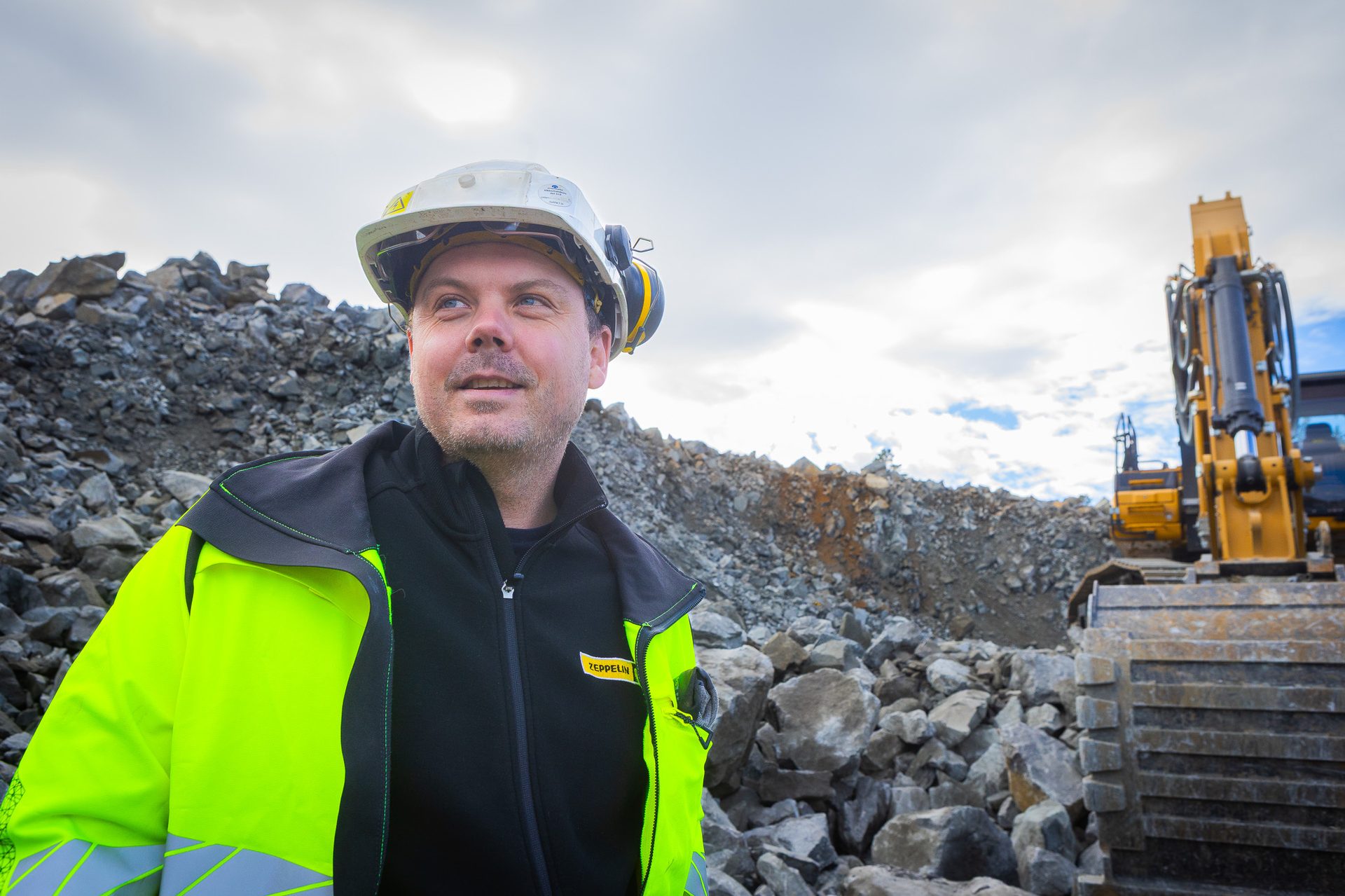 Man in hard hat and high-vis jacket looks up in a quarry with a yellow excavator and rock piles.