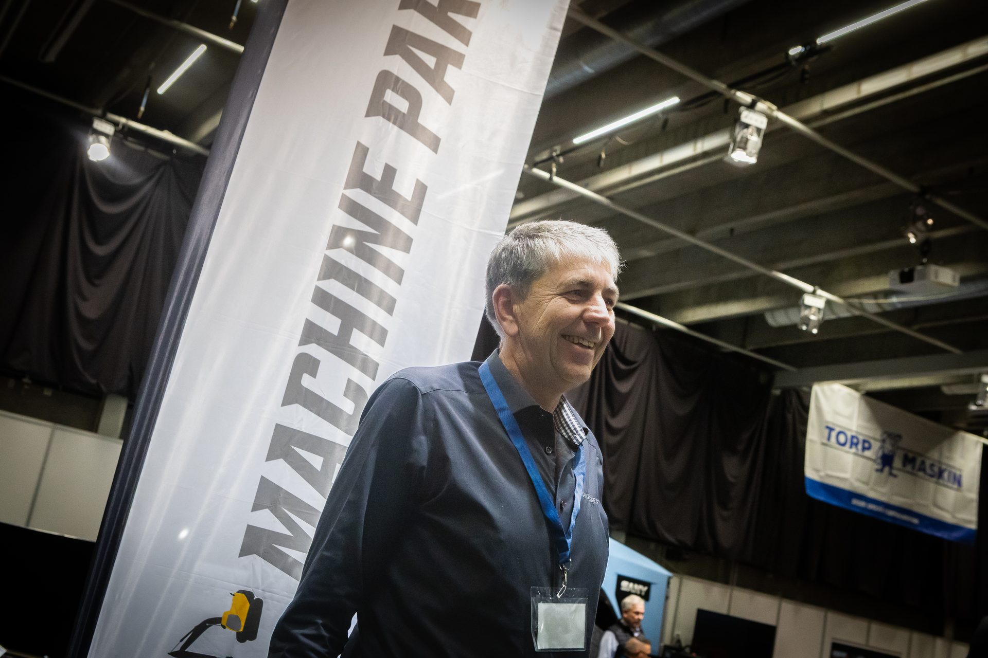 A smiling, grey-haired man with an ID badge at an exhibition next to a "MACHINE PART" banner.