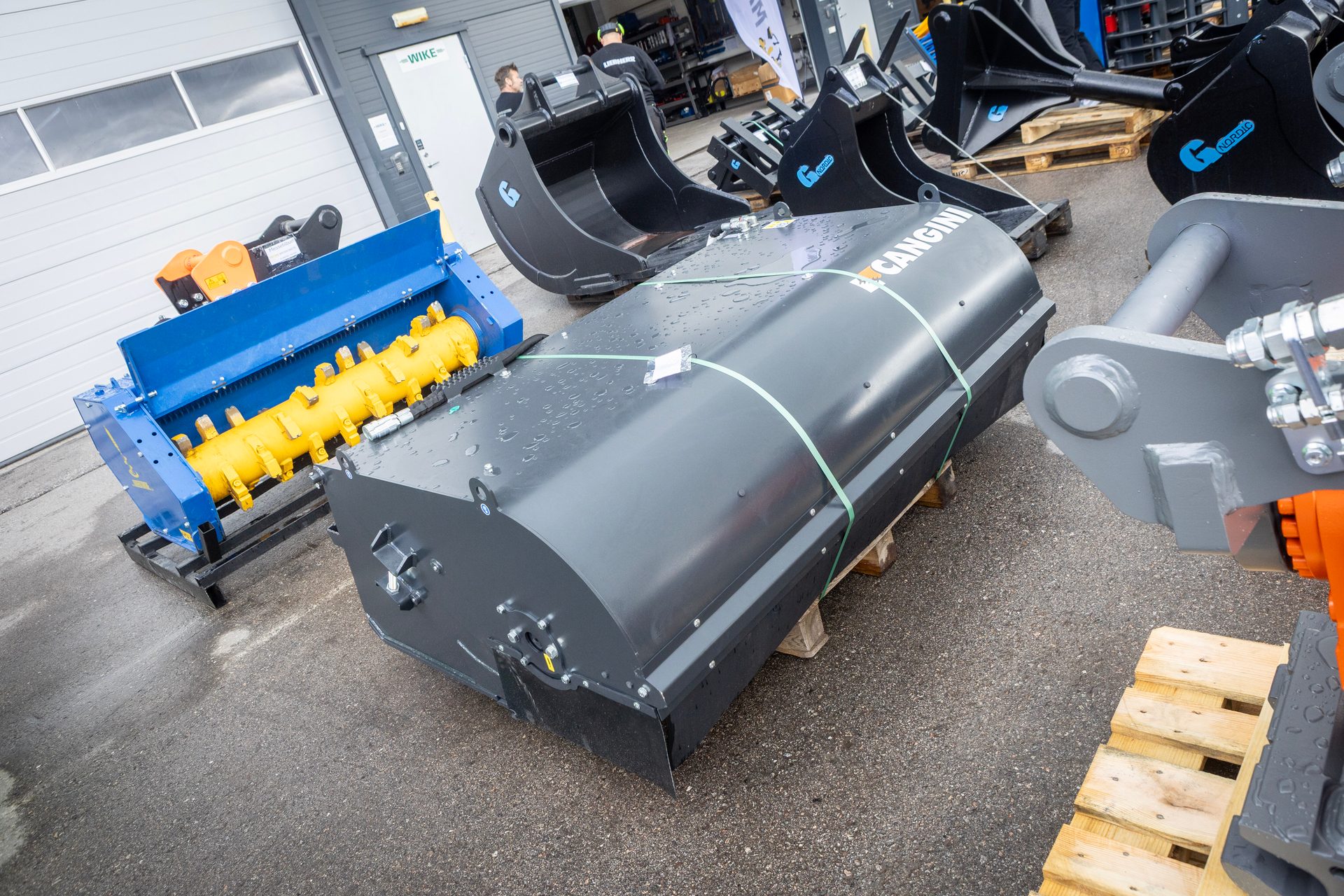 Various heavy equipment attachments including a blue mulcher, a gray sweeping attachment, and excavator buckets.