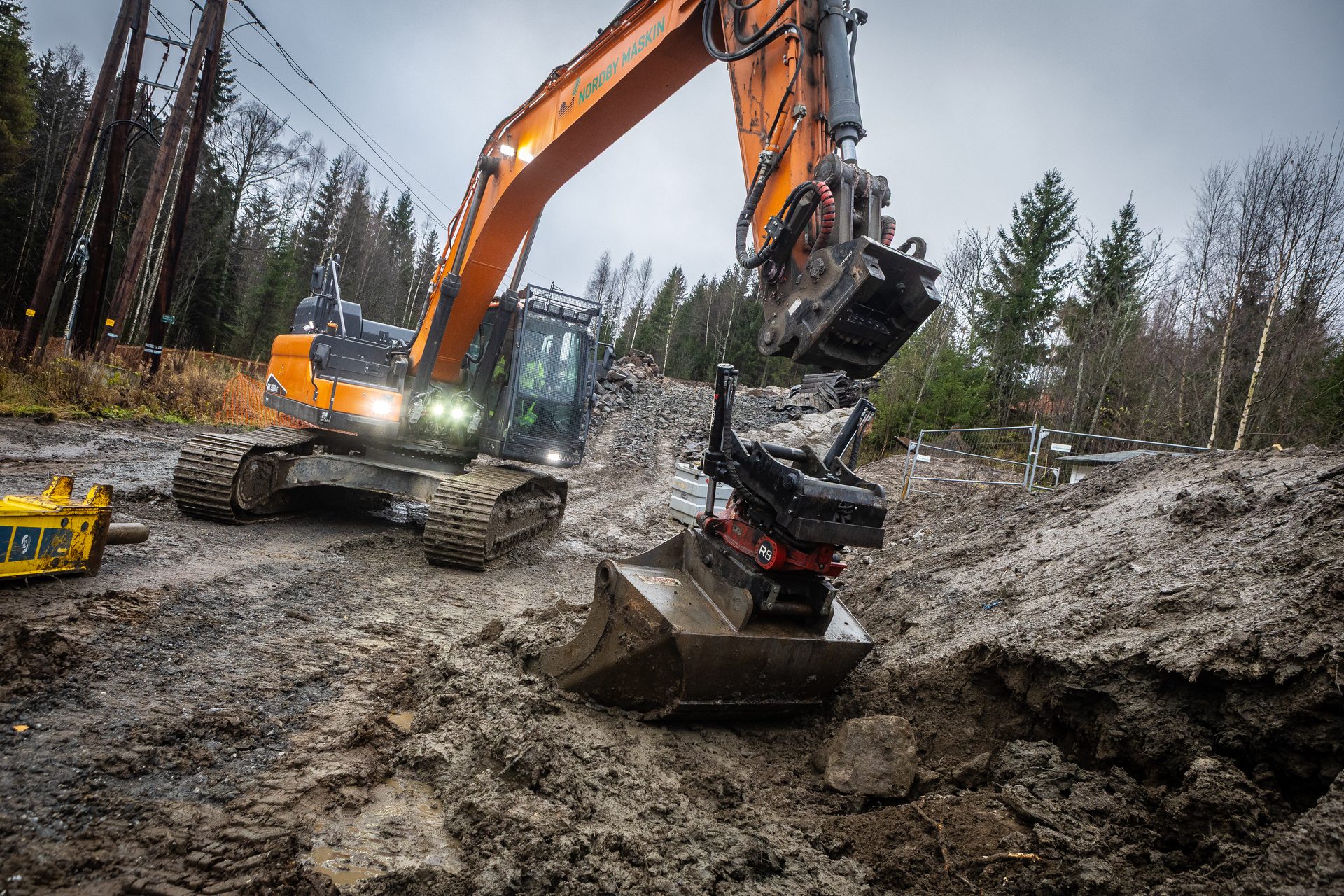 Orange excavator digs in muddy forest clearing under power lines.