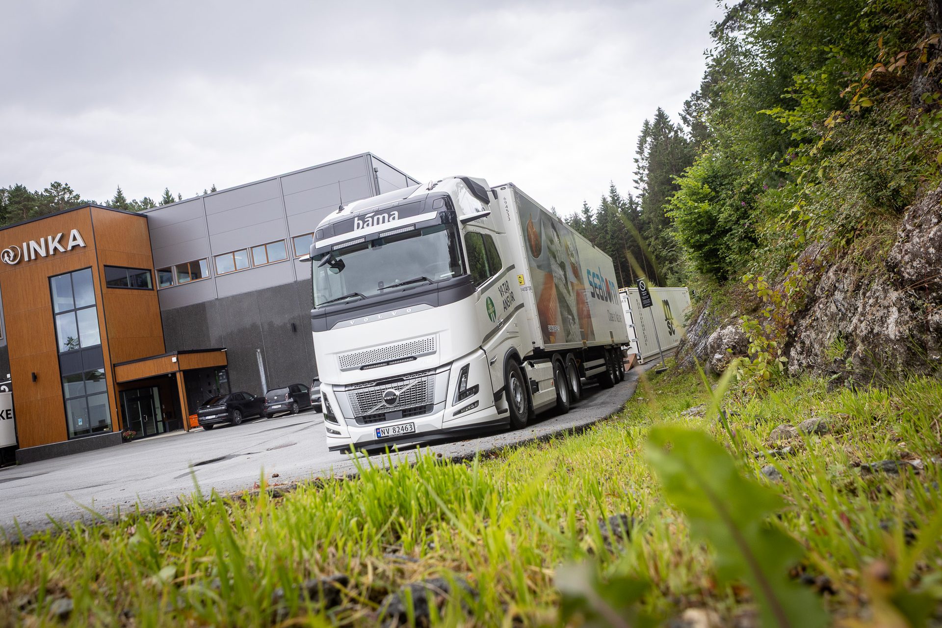 A white Volvo truck with 'bama' logo and 'SEADAN' trailer parked next to the modern 'INKA' building.