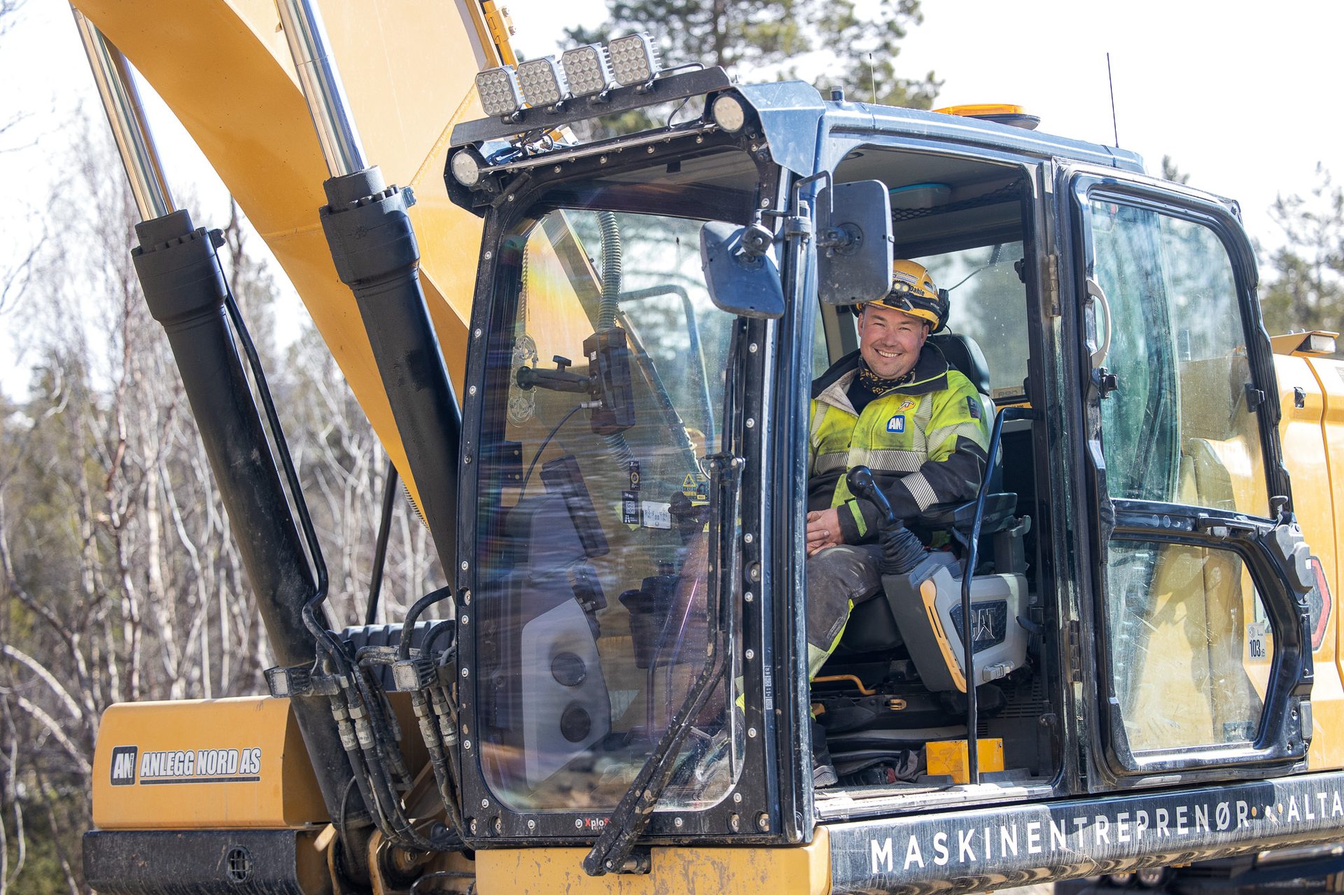 Smiling man in a yellow hard hat and safety jacket operating an excavator in a natural setting.