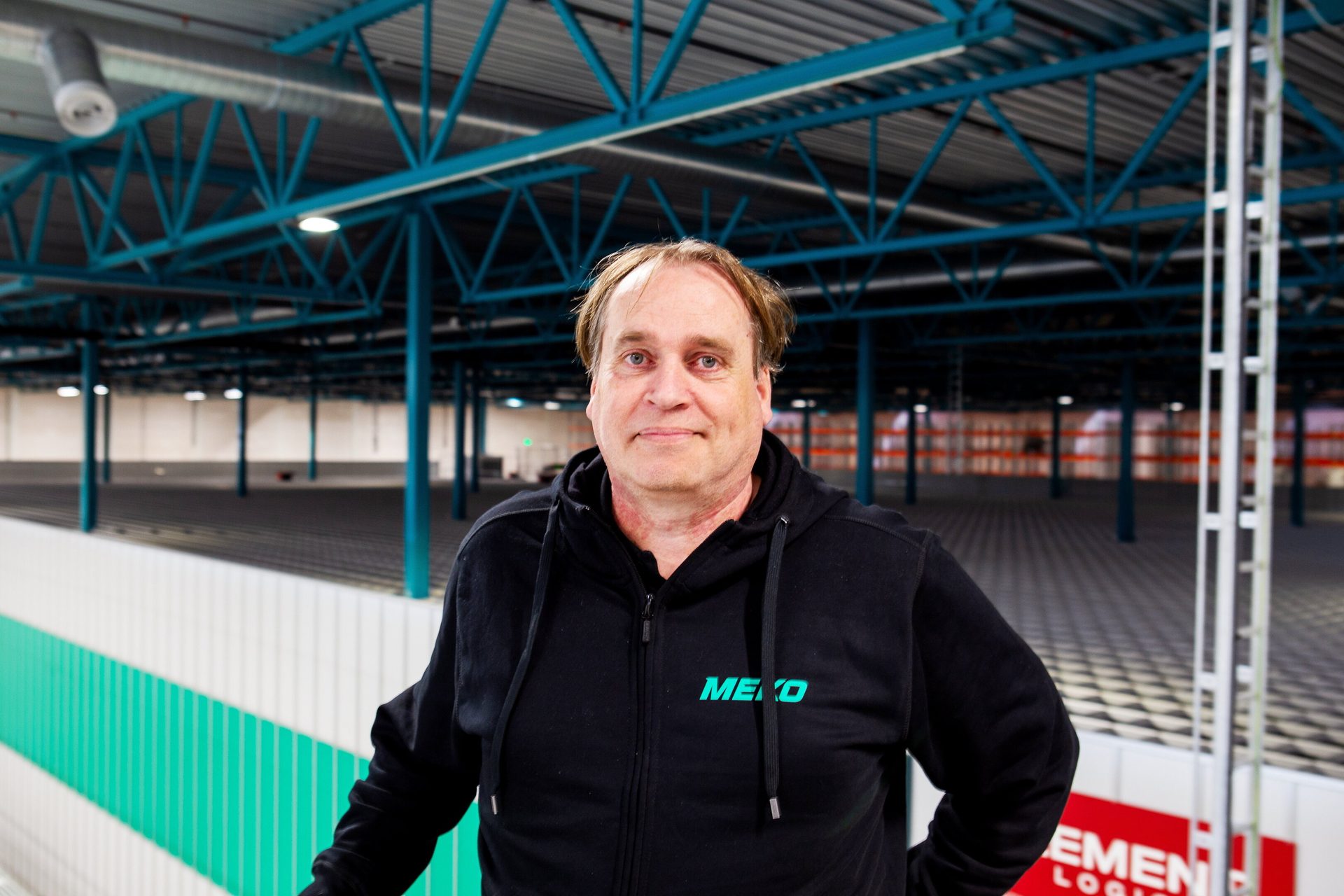A man in a black hoodie stands in an empty warehouse with a teal structural ceiling.
