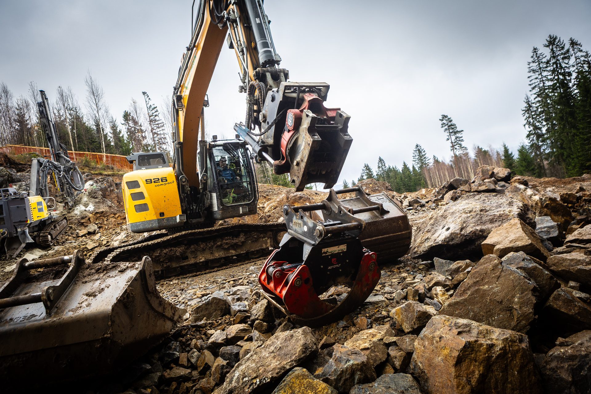 Yellow excavator with a grab attachment and a drilling rig on a rocky construction site.