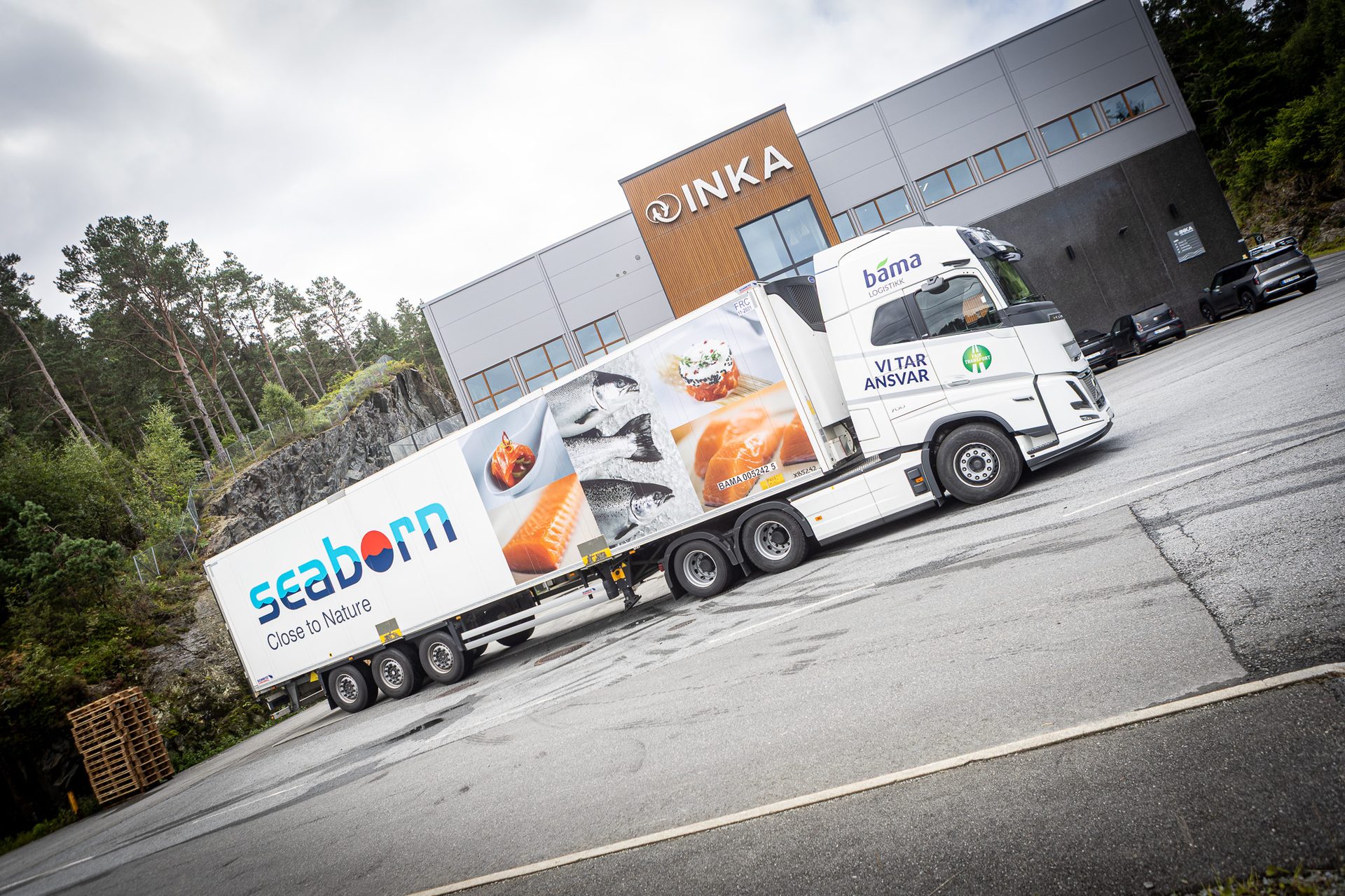White "Seaborn" truck with seafood graphics parked in front of an "INKA" building, surrounded by trees.
