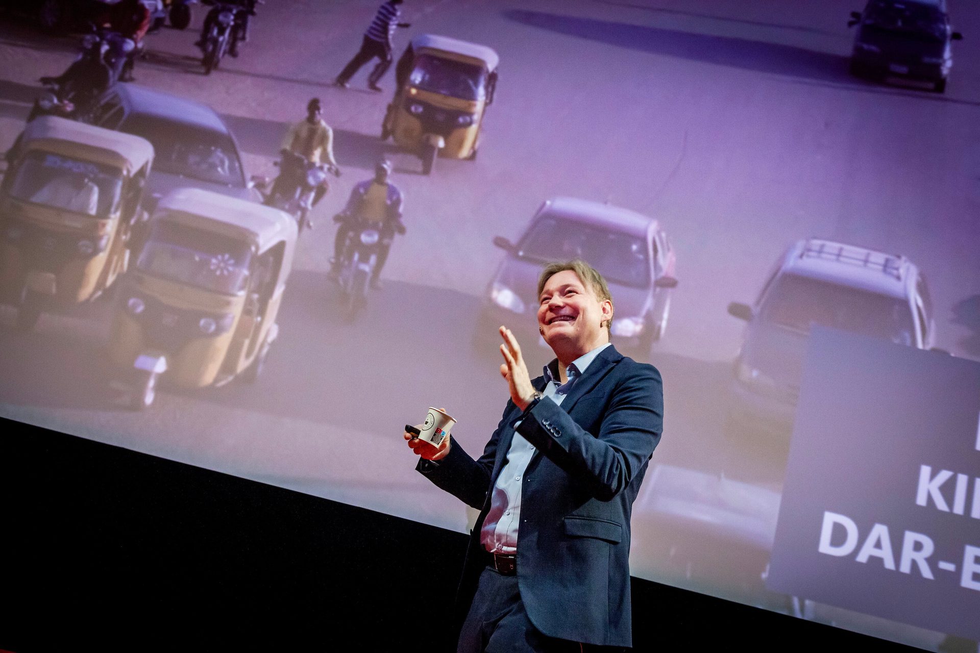 A smiling man on stage gestures, holding a cup, with a large screen projecting a busy street scene.