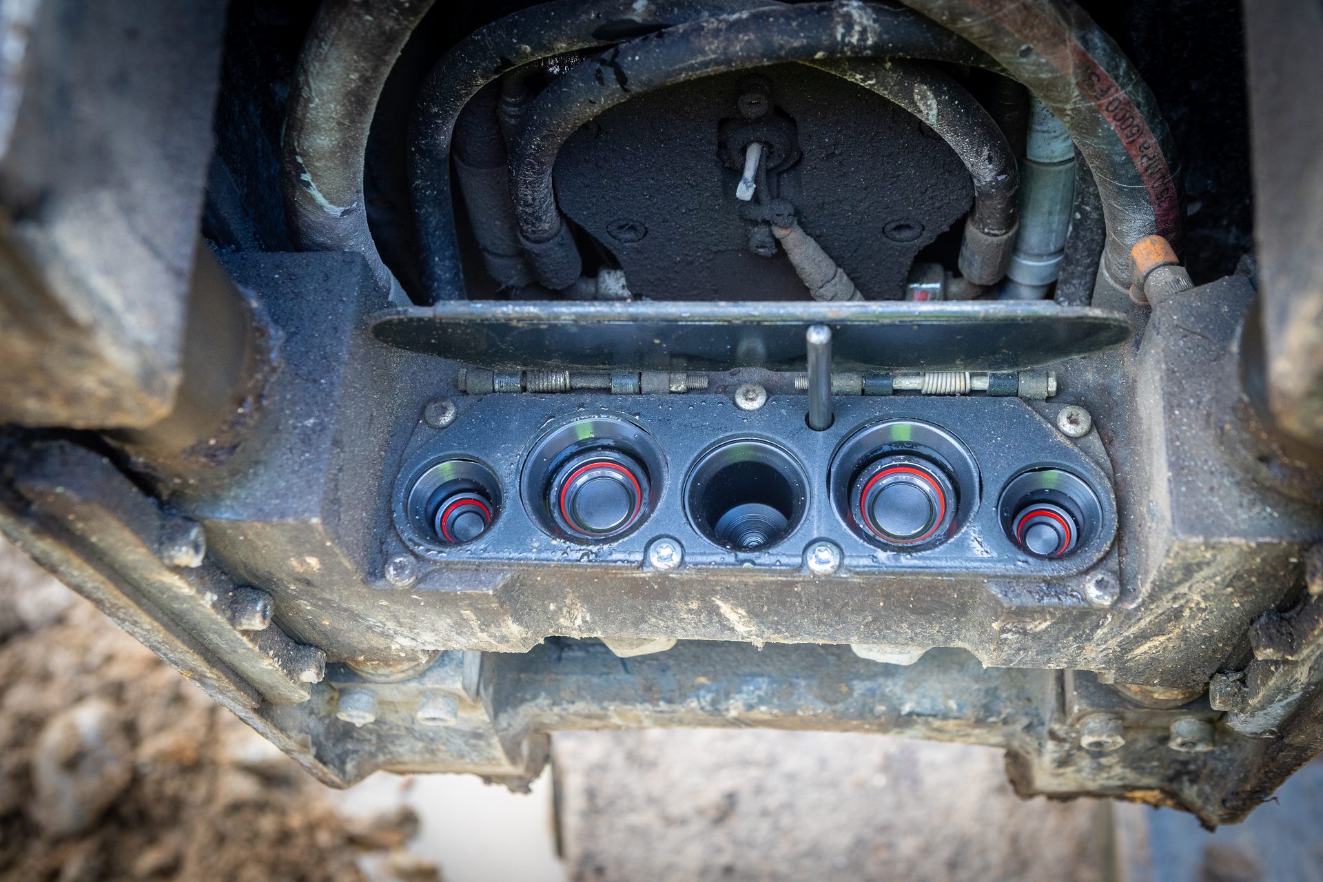 Dirty heavy machinery's hydraulic hoses and panel of circular ports, some with red rings.