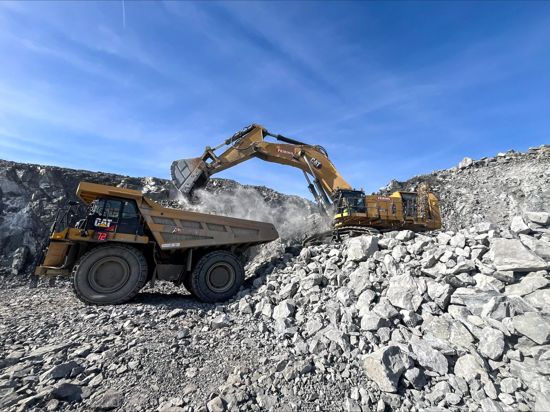 A large CAT excavator loads rocks into a dump truck in a dusty quarry operation under a blue sky.