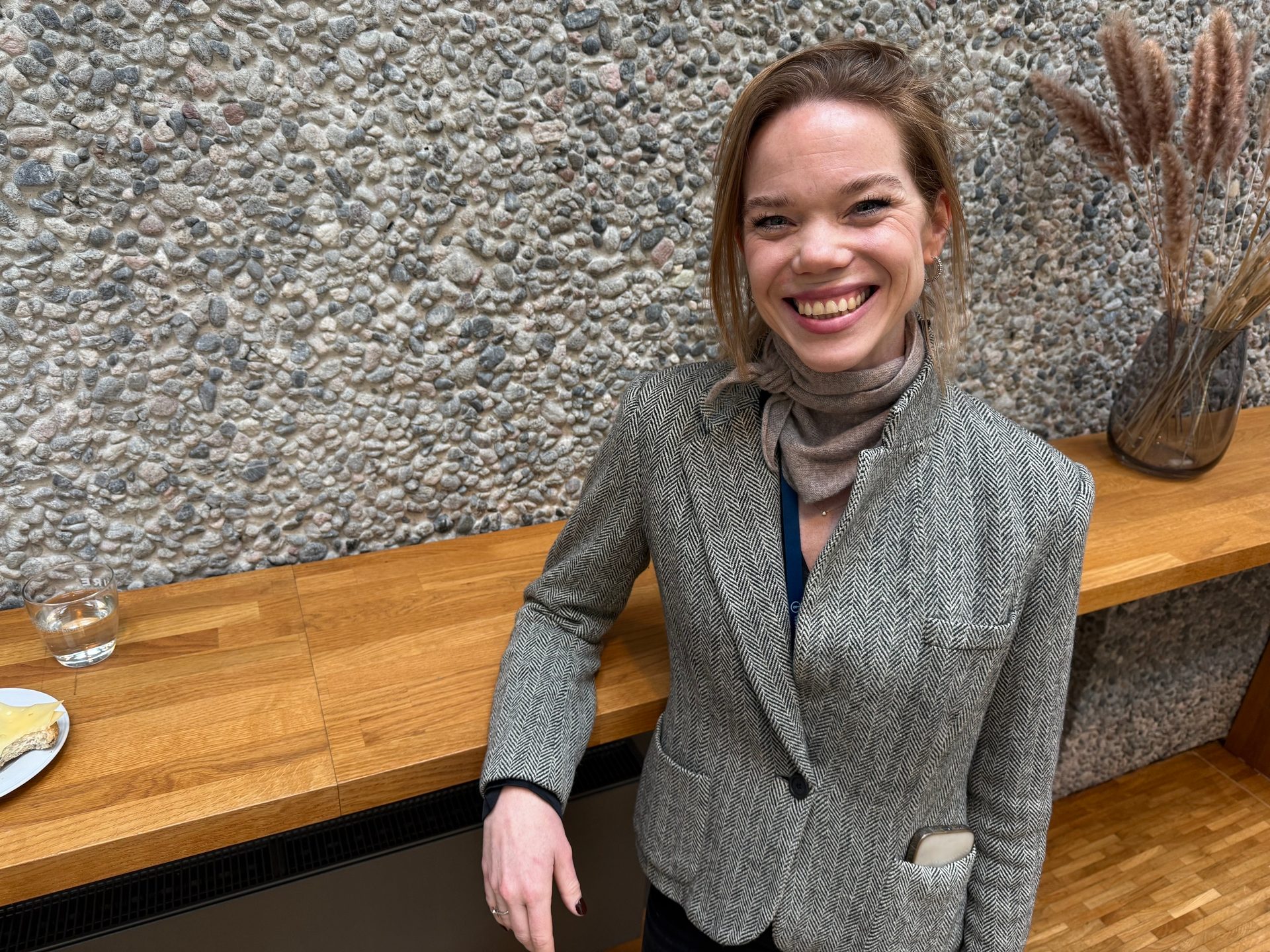 Smiling woman in a tweed jacket and scarf, leaning on a wooden counter against a stone wall.