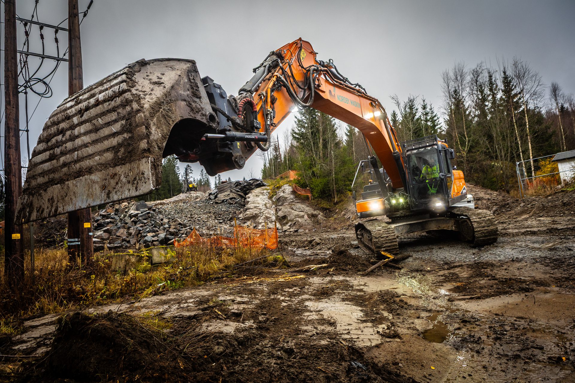 Heavy excavator with giant dirty bucket works on muddy site near power lines.