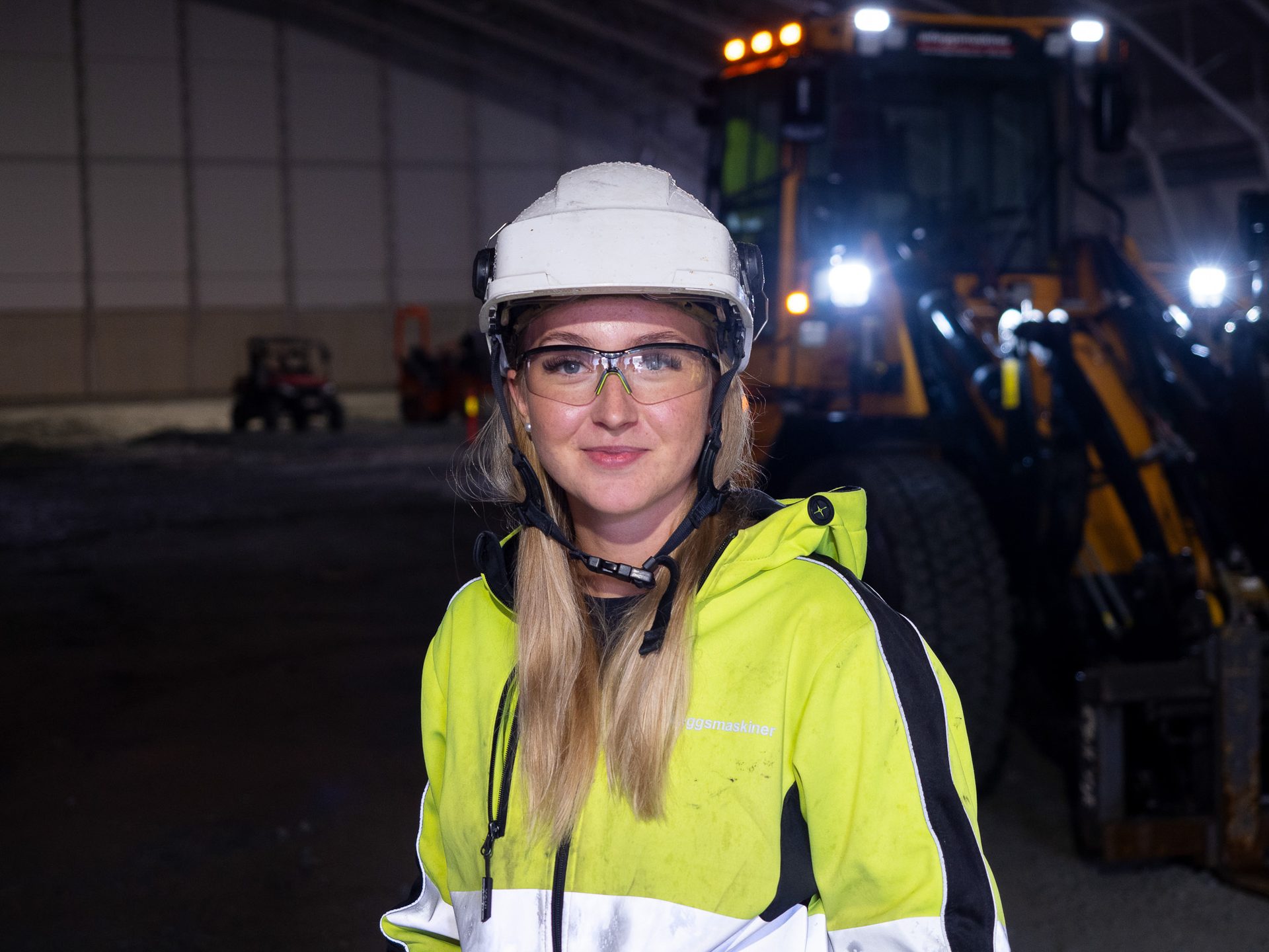 Smiling woman in hard hat, safety glasses, and yellow high-visibility jacket in industrial setting.