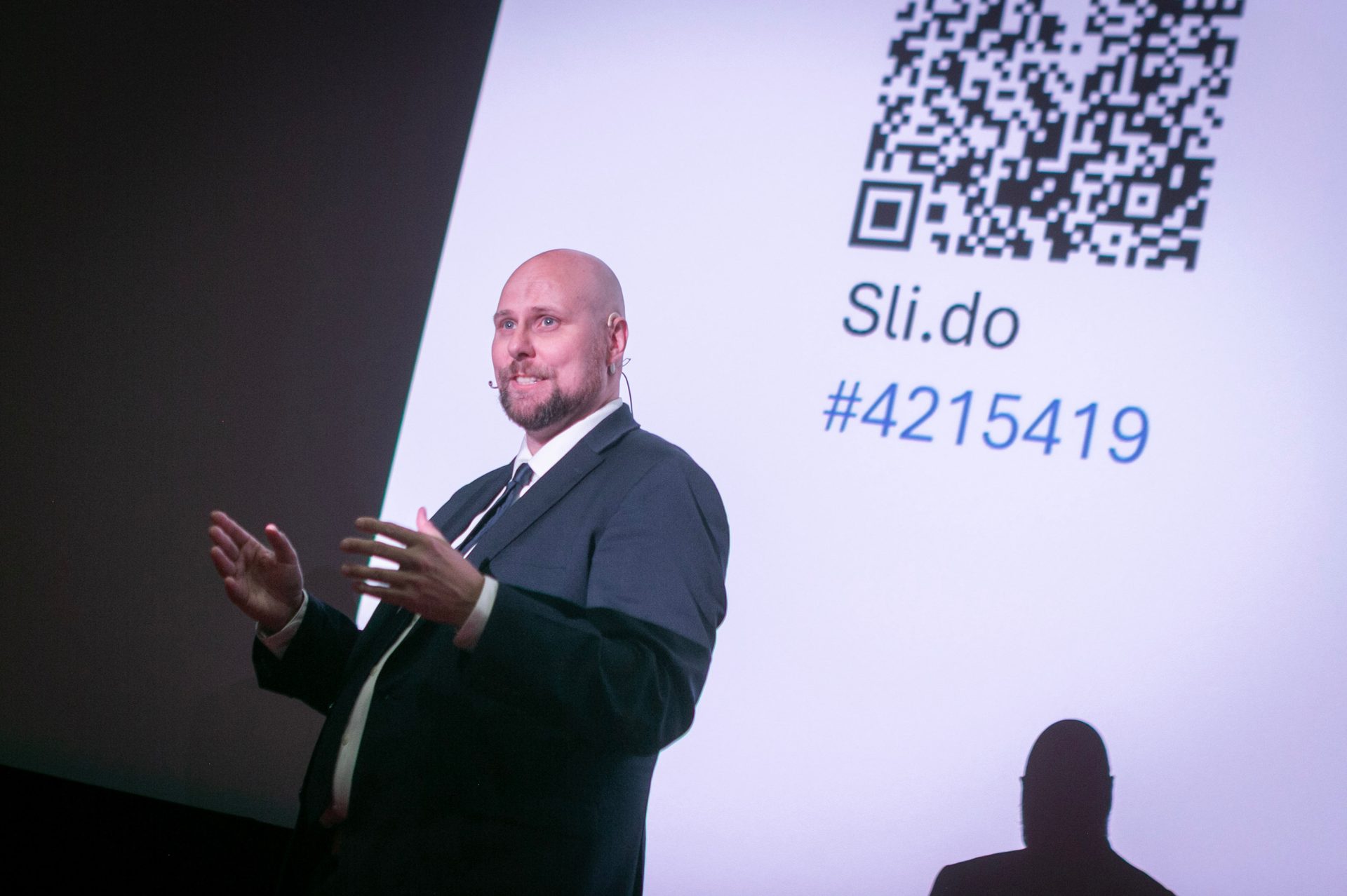 Bald man in suit presenting, shadow on screen with QR code and Sli.do #4215419.