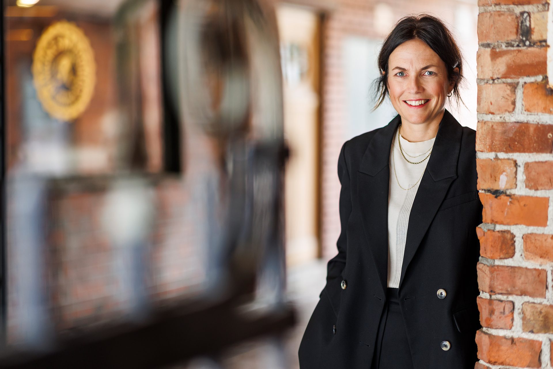 Smiling woman in a black blazer by a brick wall, blurred background.
