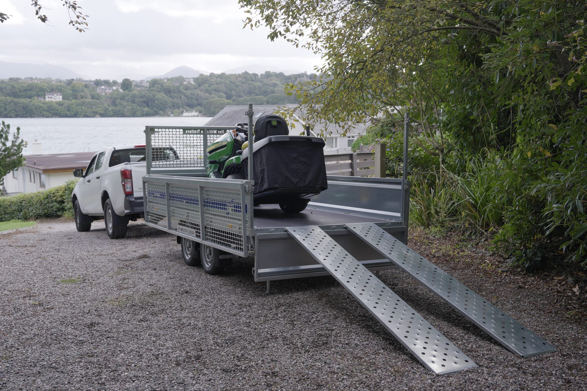 White pickup truck towing a trailer with a John Deere lawnmower and loading ramps next to a lake.