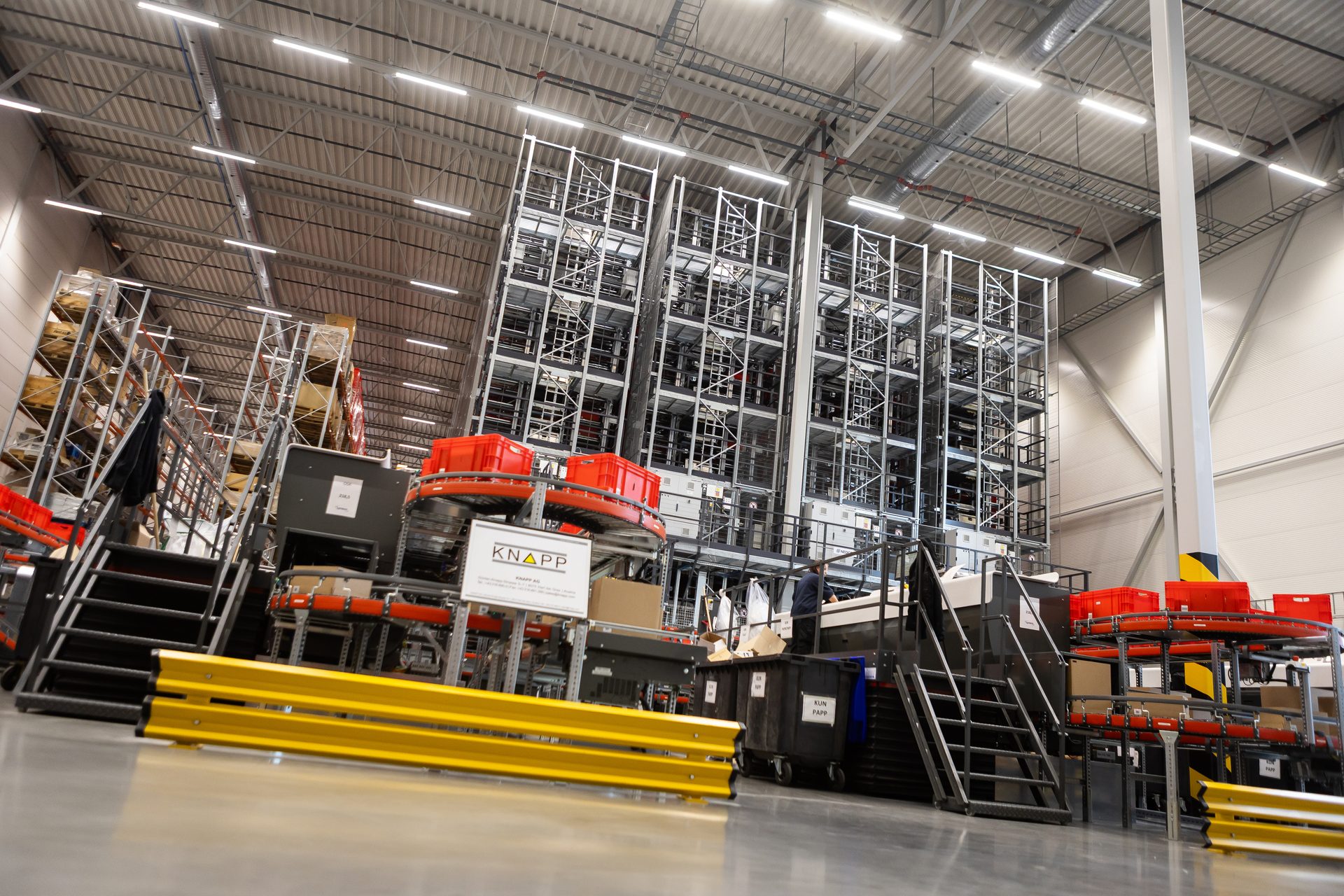 Automated warehouse with towering KNAPP storage systems, conveyor belts, and workstations for package handling.