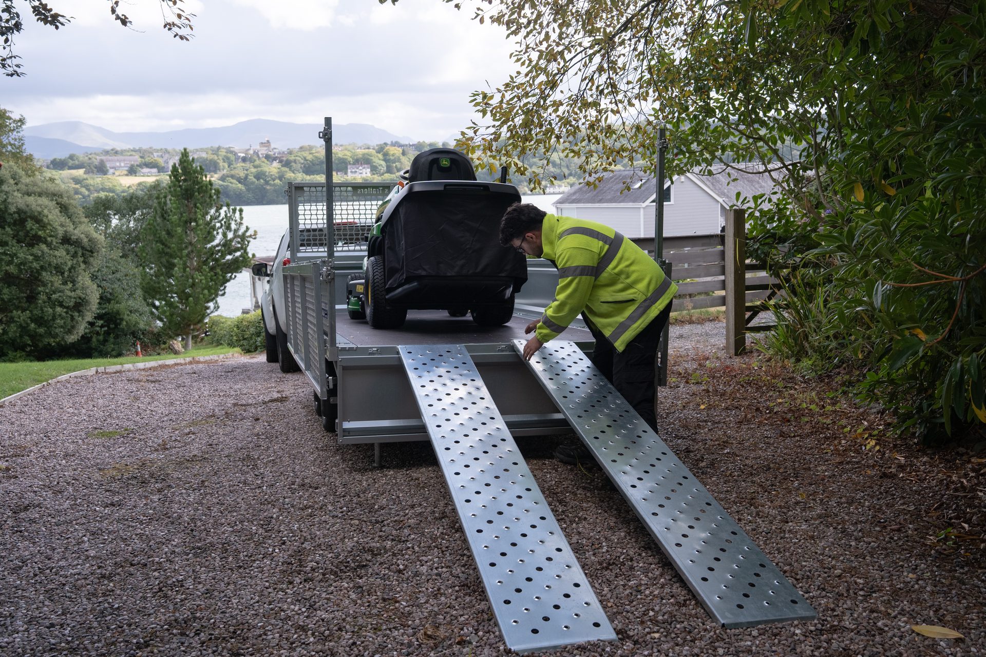 A man in a high-vis jacket setting up metal ramps behind a trailer loaded with a lawnmower.