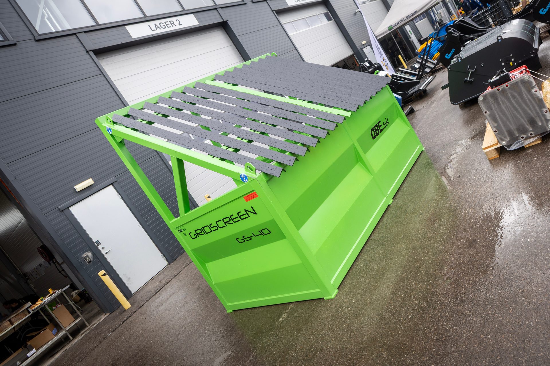 Bright green industrial grid screen with a slatted top, parked on wet asphalt outside a warehouse building.