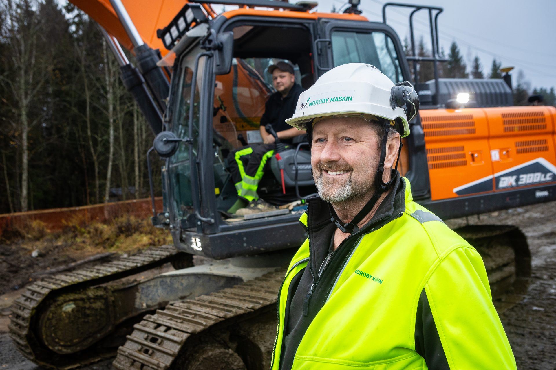 Two men, one smiling in a hard hat and safety vest, with another in an orange excavator.