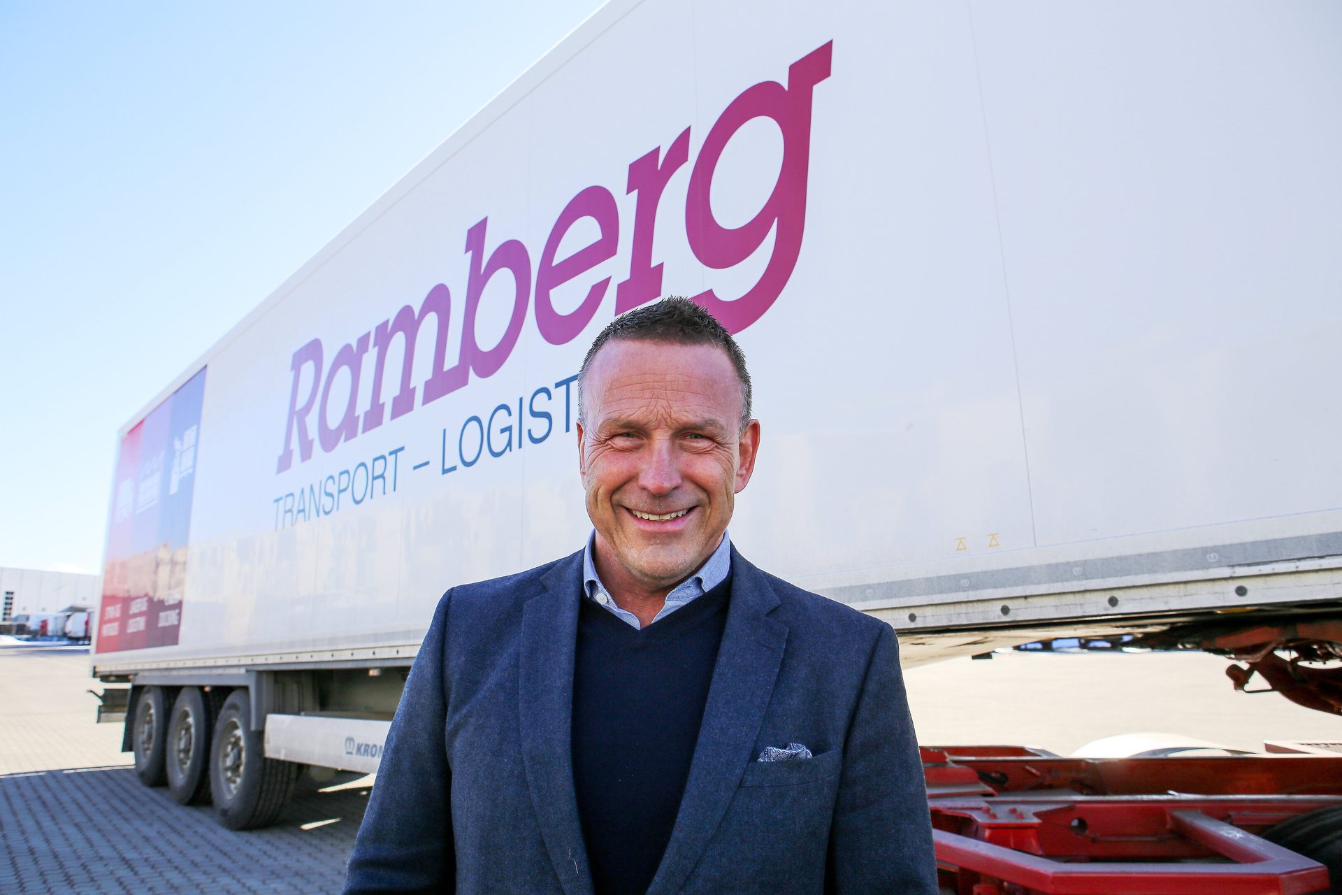 Smiling man in suit standing before a white Ramberg Transport-Logistik truck trailer on a sunny day.