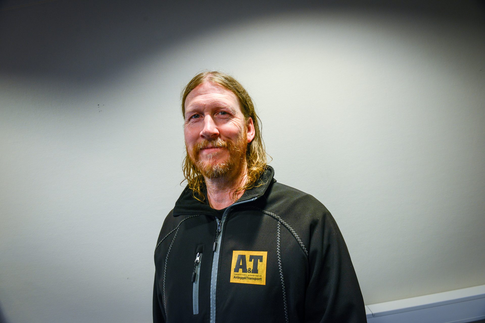 Smiling man with long hair & beard in black jacket with yellow "AT Anlegg & Transport" logo.