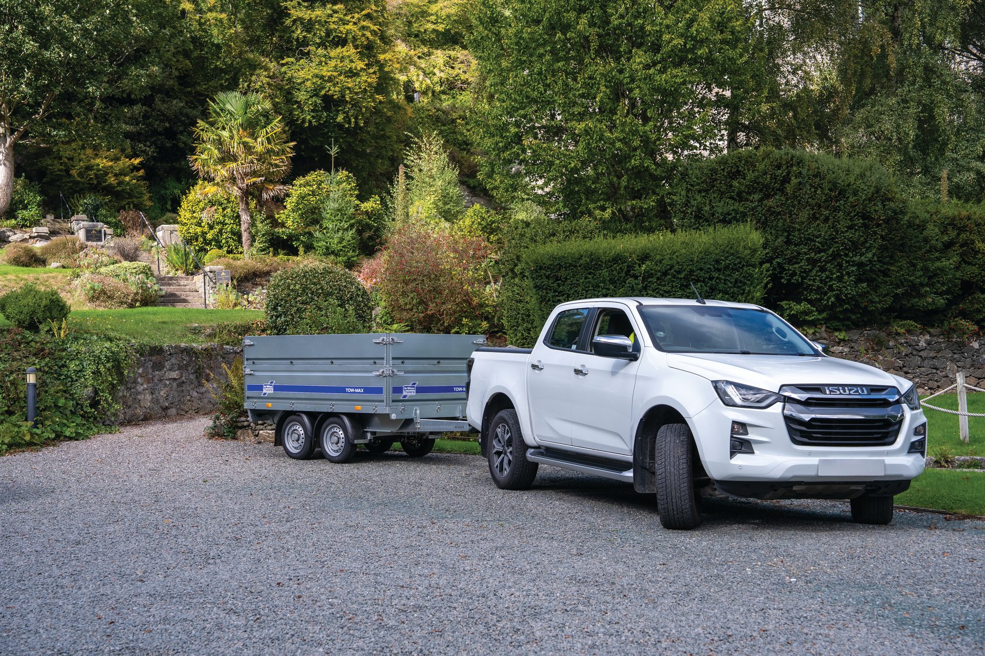 White Isuzu D-Max pickup towing a gray trailer on a gravel driveway with a lush garden.