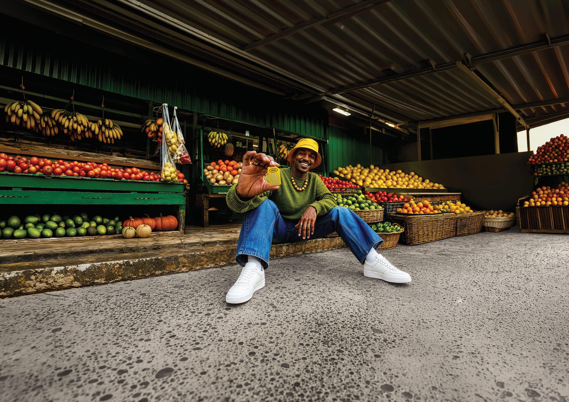 Man with a yellow hat sitting at a vibrant fruit and vegetable market, holding up a golden object.