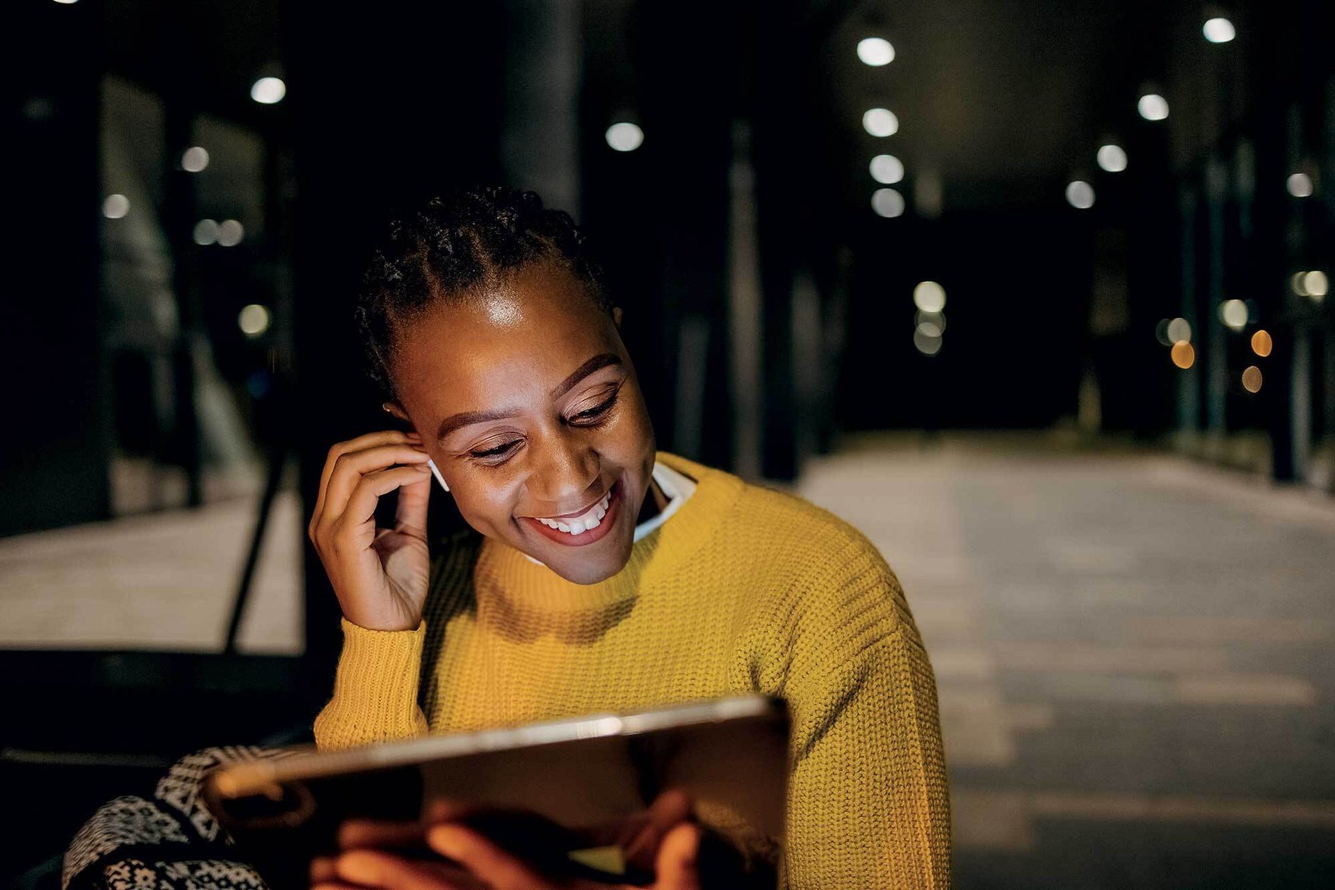 Smiling Black woman adjusting earbud while looking at a tablet at night.