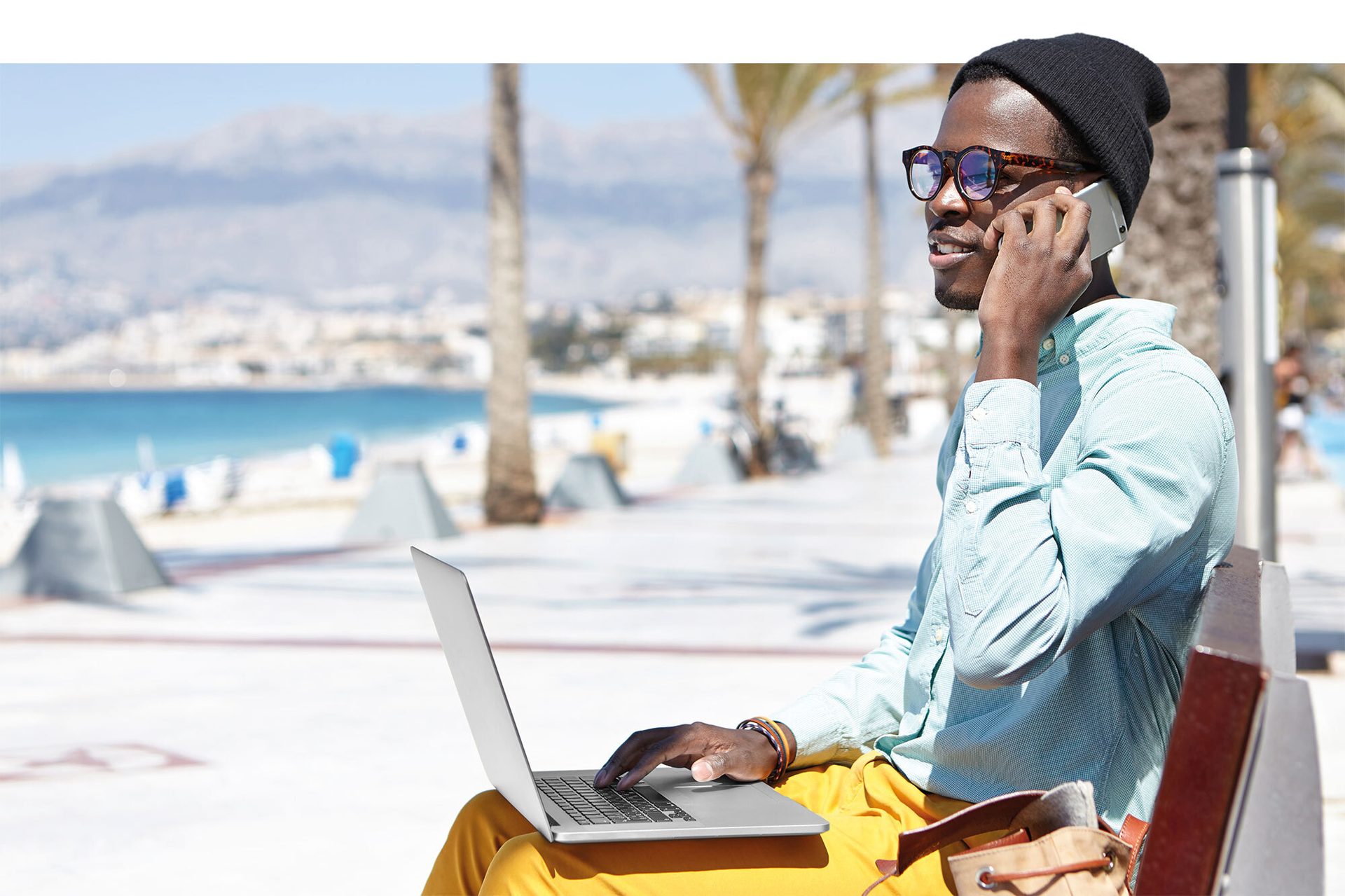 Black man on bench talking on phone, using laptop, with beach and mountains in background.