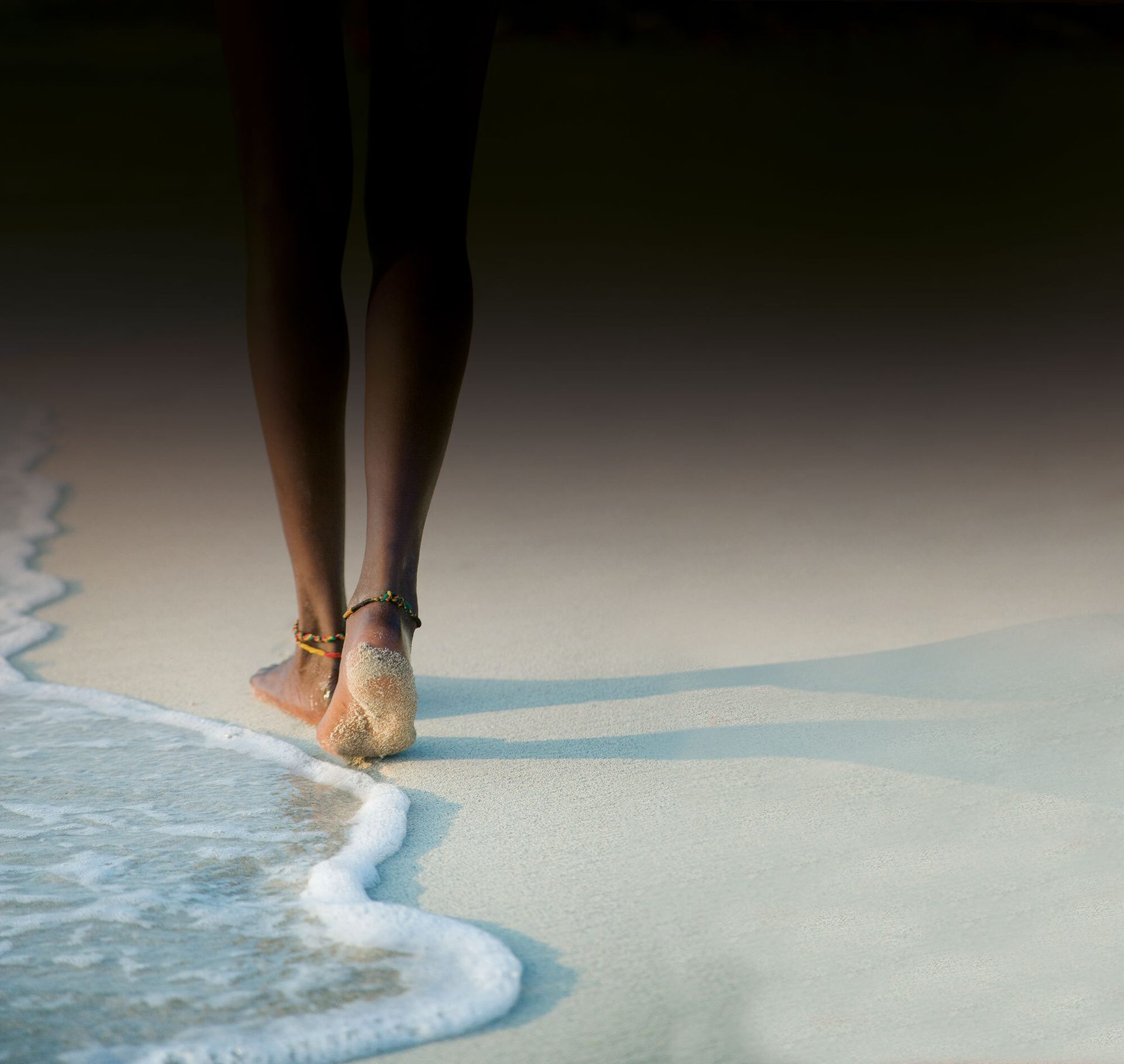 Person walking away on a sandy beach, bare feet with anklets in the ocean foam.